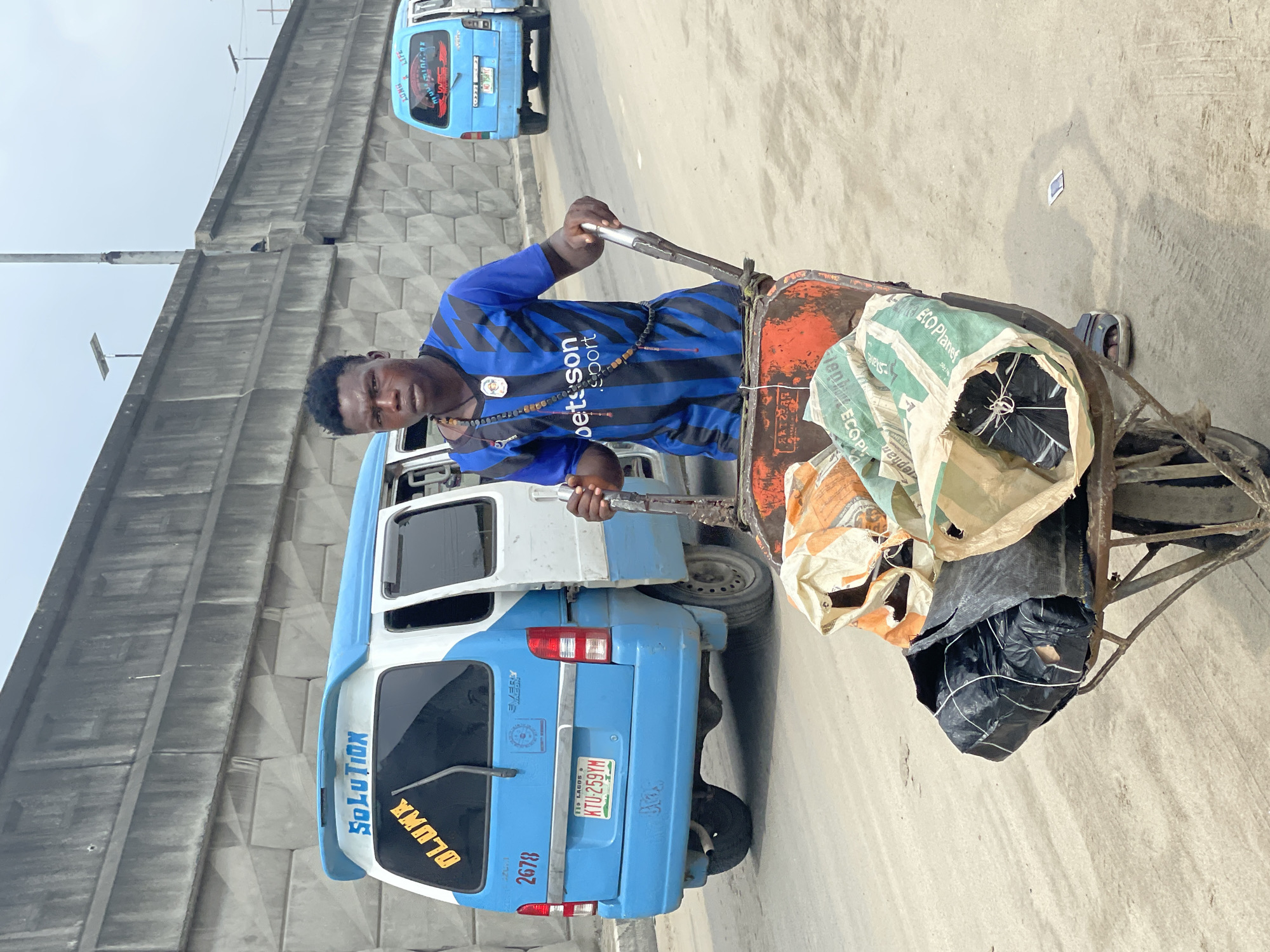 A young Black man in a blue and black striped soccer jersey and a necklace of wooden beads pushes a wheelbarrow loaded with sacks on a sandy road in Lekki, Nigeria. The wheelbarrow, made of orange metal, contains several large sacks, some open and revealing black plastic-wrapped packages. In the background, a light blue and white commercial van with the word "SOLUTION" written on its rear spoiler is parked, its license plate reading "KTU-259YM". Another light blue van is visible further down the road. A concrete wall with a textured, angular pattern forms a backdrop behind the vehicles and the man. The sky is overcast.