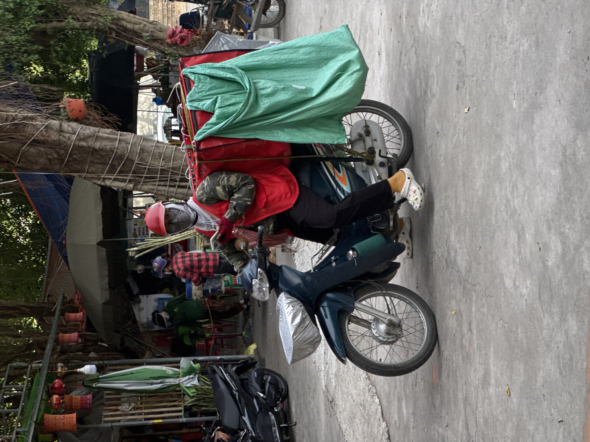 A person wearing a red helmet, a red vest over a camouflage shirt, and dark pants is riding a teal motorcycle. The motorcycle has a silver bag in the front basket and a large green bag attached to the back. The person is holding a red object, possibly a phone, in their right hand. The scene appears to be a street market in Hoa Lư Ward, Vietnam, with stalls, umbrellas, and other motorcycles visible in the background. The ground is paved with concrete.
