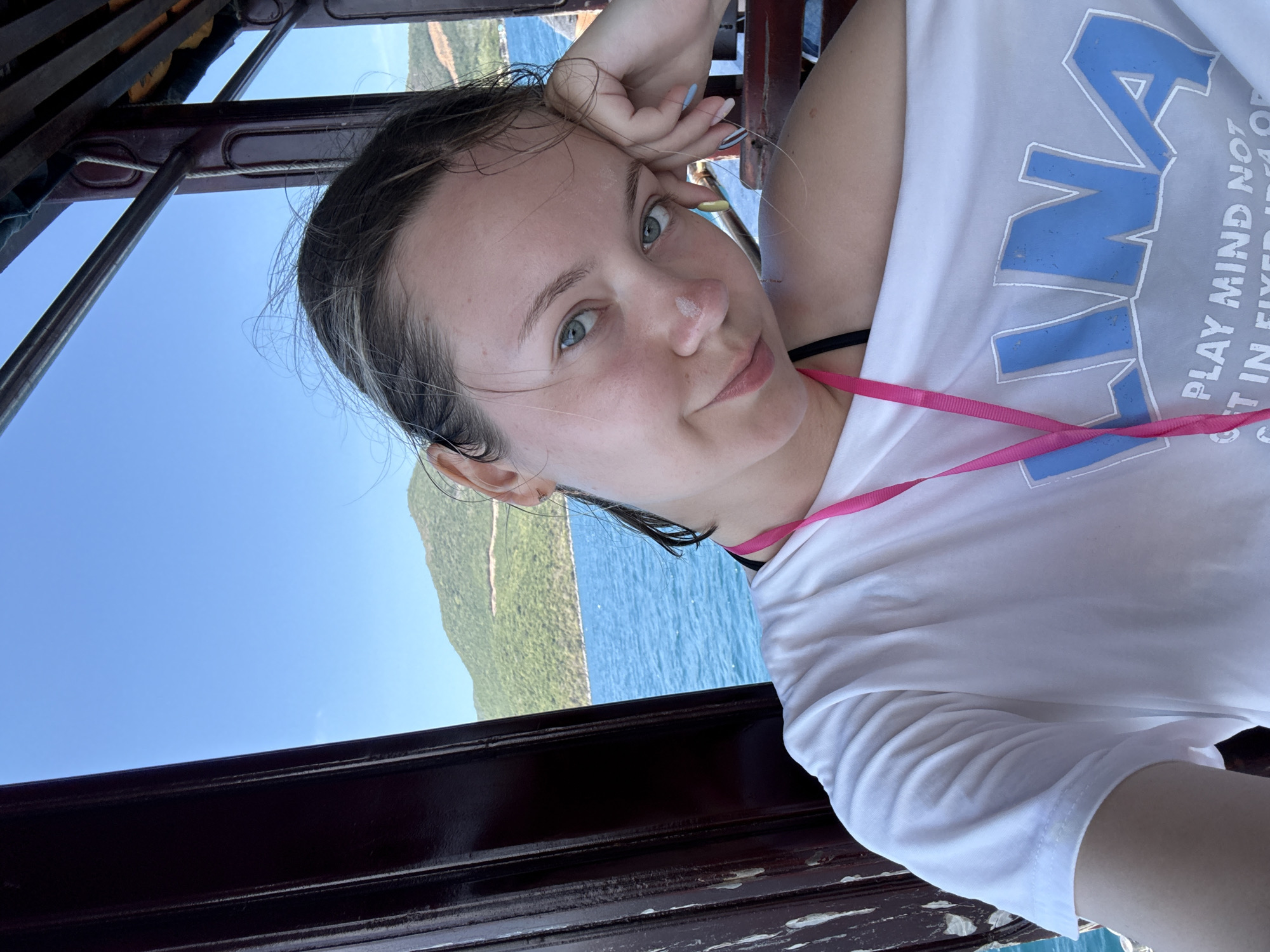 A woman with wet hair and green eyes, wearing a white t-shirt with blue lettering and a pink lanyard, poses for a selfie. She is on a boat, evidenced by a dark wooden railing in the foreground. In the background, there is a view of the ocean, a small island, and a clear blue sky. Based on the available information, the location is Nha Trang Ward, Vietnam.