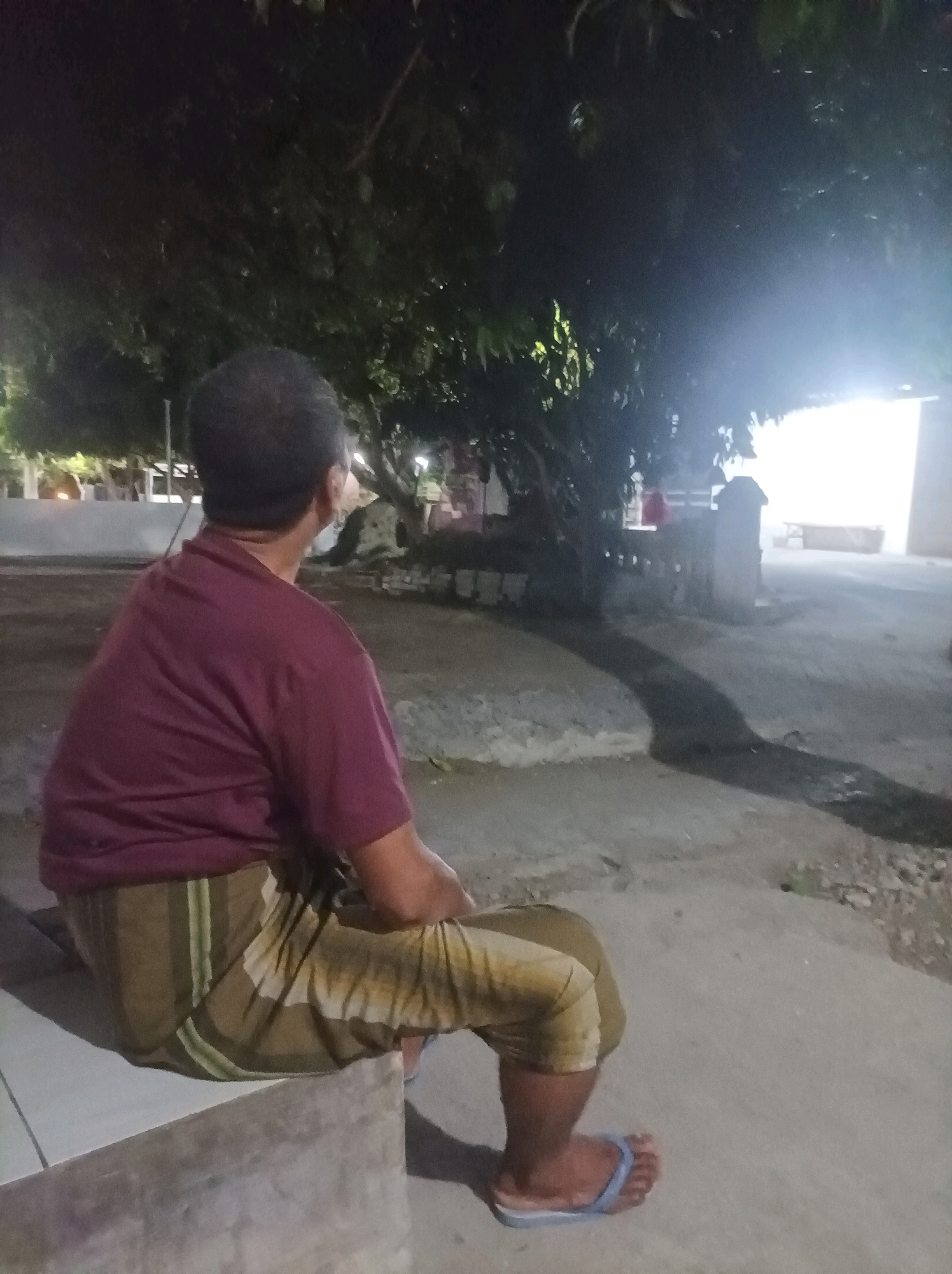 A man, viewed from behind, sits on a concrete ledge outside at night. He is wearing a dark maroon t-shirt and a patterned sarong in shades of brown and green. He is looking to the right, towards a brightly lit area. The background consists of trees and a building with a bright light emanating from it. The ground is paved with concrete and dirt. The location is identified as Canga'an, Indonesia.