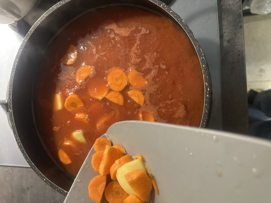 A top-down indoor view captures a cooking process in a kitchen environment. A dark-colored cooking pot with a speckled rim rests on a stove surface, containing a simmering red-orange liquid with numerous round, orange carrot slices and lighter, irregular vegetable chunks. Steam is visible rising from the pot's contents. A light grey, flat scoop, holding additional chopped orange carrot slices and lighter-colored vegetable peelings, is positioned above the left side of the pot, indicating the imminent transfer of these ingredients into the pot. The background shows blurred portions of a countertop or stove and a dark, indistinct object, possibly a person's leg, at the very bottom. The specific location within Tbilisi, Georgia, cannot be determined from the image.