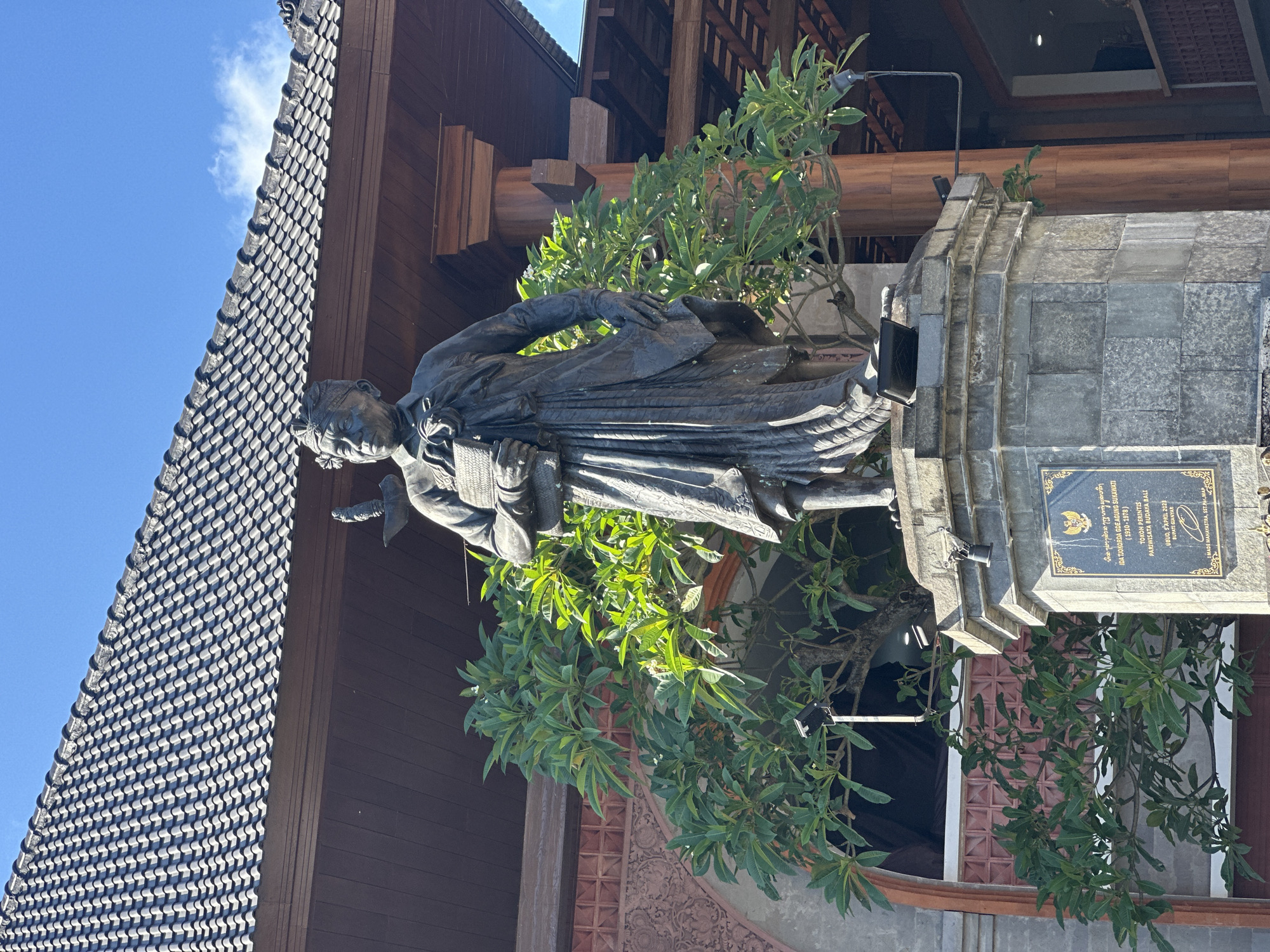 A bronze statue of a man stands on a stone pedestal in front of a building with a dark, tiled roof and wooden beams. The statue depicts a figure dressed in traditional Balinese clothing, with a head covering, and holding a book or scroll. The man has a stern expression. A lush green tree with broad leaves grows behind and to the left of the statue. A plaque with text and an emblem is affixed to the pedestal. The background shows a clear blue sky with a few wispy clouds. The scene appears to be located in Ubud District, Indonesia, as indicated by the plaque which states "TOKOH PERINTIS PARIWISATA BUDHA BALI" and "UBUR, 5 APRIL 2023, BUPATI GIANYAR." The statue is likely of Ida Tjokorda Goeang Soekawati, a key figure in the development of tourism in Bali.