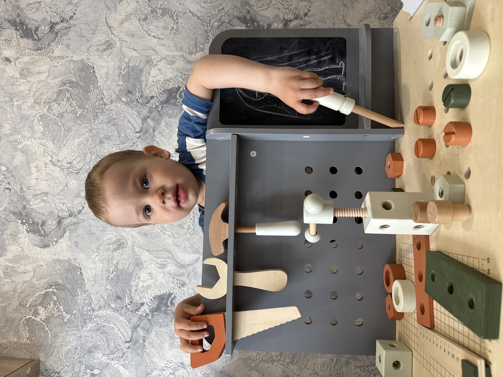 A young child wearing a blue and white striped shirt is playing with a wooden toy workbench. The workbench is gray and wood-colored, featuring various wooden tools, screws, and a chalkboard. The child is writing on the chalkboard with a wooden tool. The background is a textured, light gray wall. The image seems to be taken indoors, and no specific indicators point to Rusanovo, Russia.