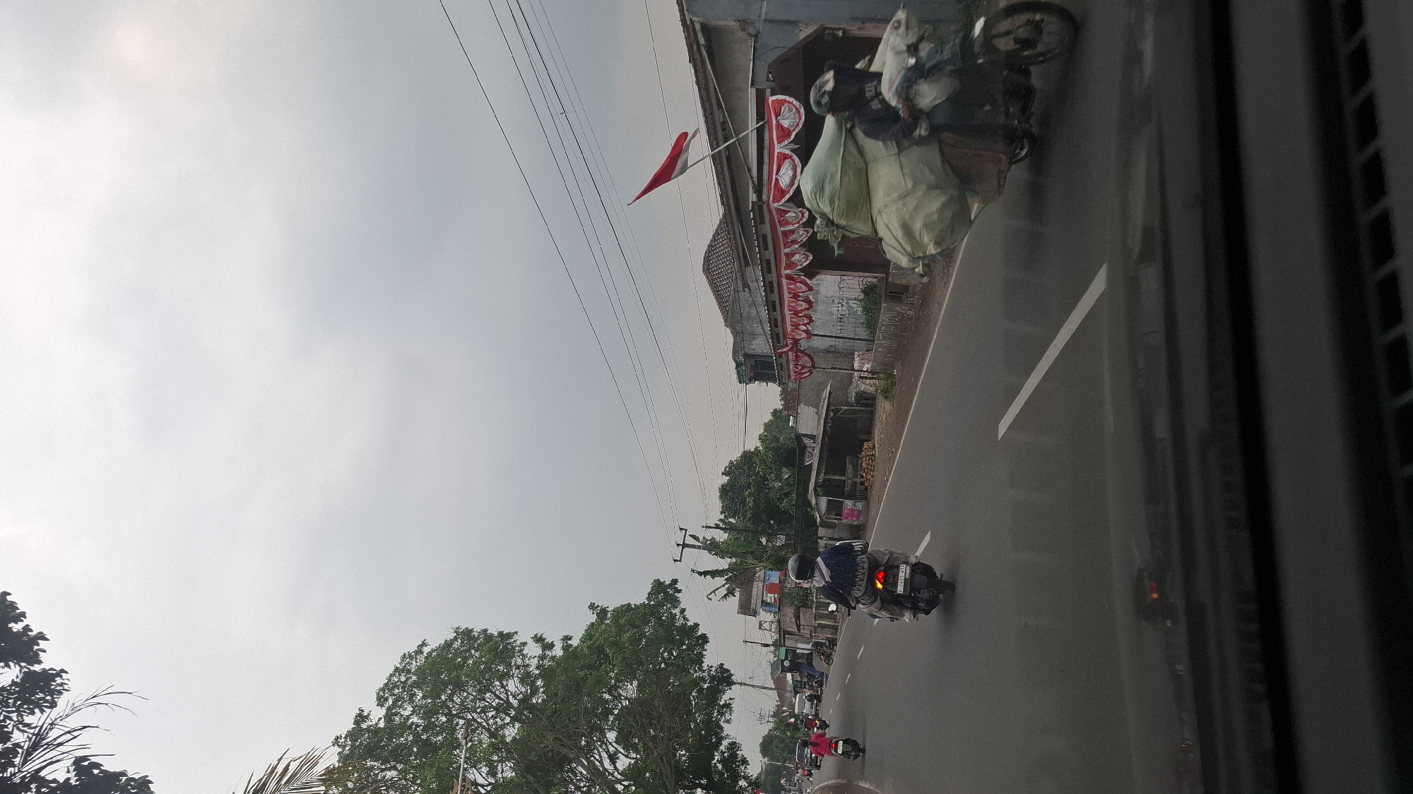 This image depicts a street scene in Cilaku, Indonesia, under an overcast sky. A two-lane road runs diagonally across the frame, with vehicles traveling in both directions. On the right side of the road, a motorcycle is heavily laden with large, light-colored sacks, the rider wearing a dark helmet. Further down the road, another motorcycle carries a person with a backpack. On the left side of the road, there are several motorcycles in motion, one with a rider in a pink shirt. Along the roadside, there are buildings with tiled roofs, some adorned with red and white decorations and an Indonesian flag prominently displayed. Trees line the left side of the road, and power lines crisscross the sky. The foreground shows the dashboard and windshield of a vehicle, suggesting the image was taken from inside a car.