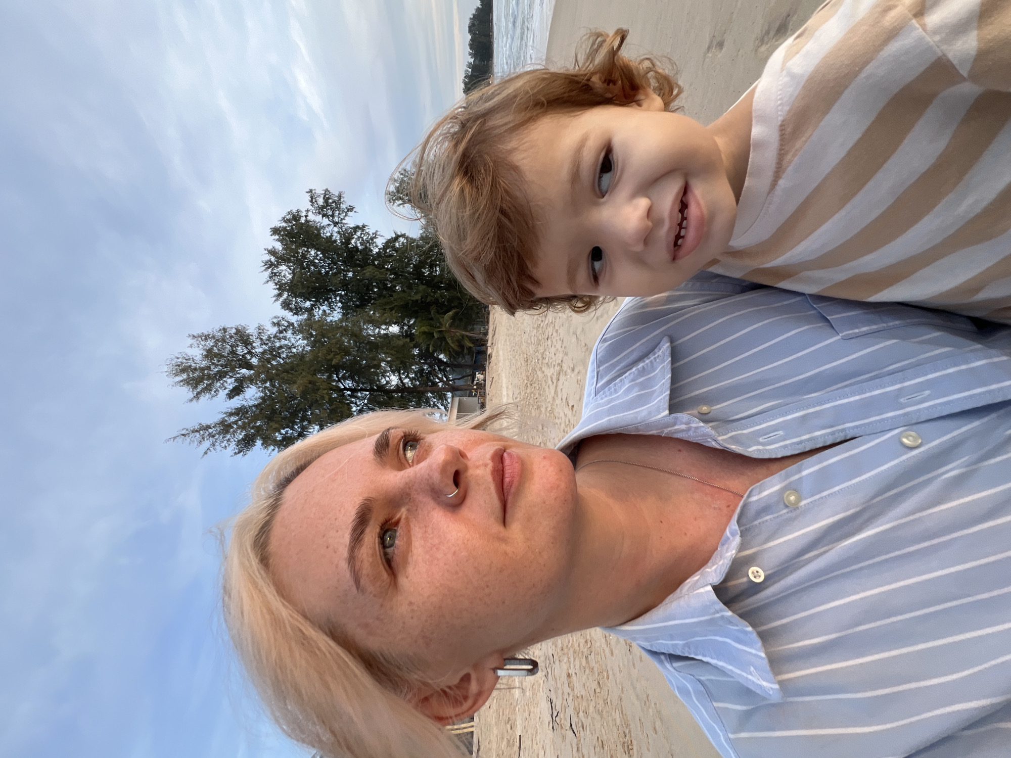 A woman with blonde hair and freckles is holding a young child on a sandy beach. The woman is wearing a light blue and white striped button-down shirt and a silver necklace. She has a hoop earring in her left ear and a nose ring. The child has curly brown hair and is wearing a beige and white striped shirt. They are both looking away from the camera, towards the ocean. The background shows trees, the ocean, and a cloudy sky. The image appears to have been taken during the late afternoon or early evening, given the lighting. The location is identified as Choeng Thale, Thailand, a coastal town known for its beaches.
