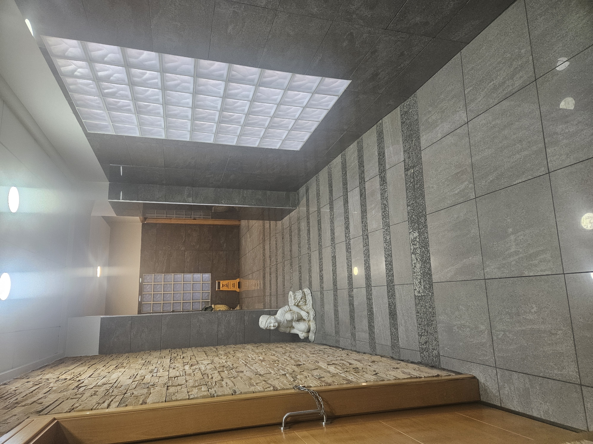 The image shows the interior of the Santuario del Rosario, a church in Hellín, Spain. The view is from near floor level, angled upward towards the ceiling. The space includes tile walls with decorative vertical stripes, and a ceiling with a translucent grid pattern in the center. A white sculpture of a seated figure is positioned along one wall. The floor is a mixture of stone-like paving and a wood-tone surface, with a chain barrier visible. An alcove with a window-like structure is visible in the background. The lighting is a mixture of natural and artificial, with bright areas and shadows.