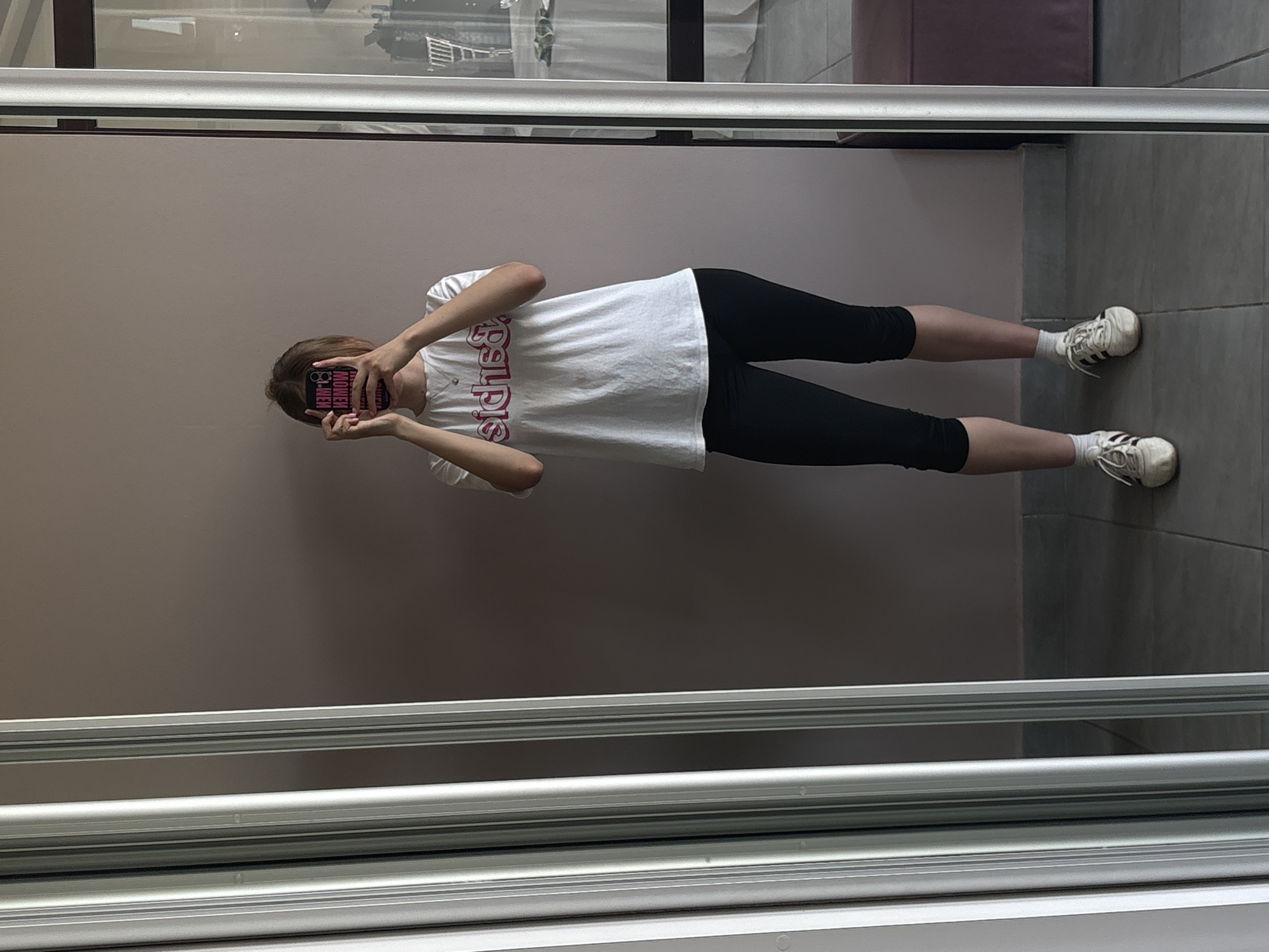 A young individual stands indoors, likely within a residential building. They are using a mobile phone with a pink and black patterned case to take a picture of themselves in a mirror. The individual wears a white t-shirt with red lettering, black leggings, white socks, and white Adidas sneakers. The background appears to be a wall painted in a light brown shade, with grey floor tiles in the reflection. The scene is captured through a mirror with a silver frame, situated indoors. Based on the prompt, the location is in Нижние Муллы, Russia.