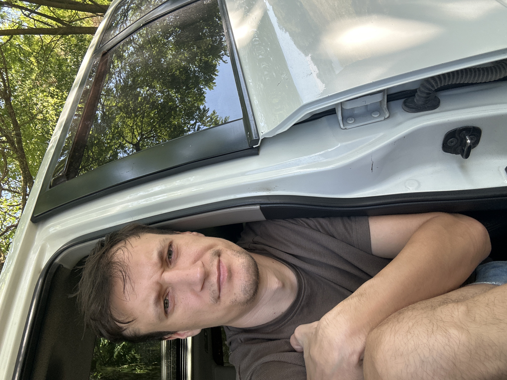 In Makiv, Ukraine, a fair-skinned man with short, dark brown hair and a light beard is sitting in the open driver's side door of a white vehicle. He is looking directly at the camera with a neutral expression. He is wearing a dark brown t-shirt and blue shorts. The interior of the car is visible behind him, with the driver's seat and a portion of the dashboard. Sunlight filters through the trees in the background, casting shadows on the car's exterior. The car appears to be parked outdoors on a dirt or gravel surface, surrounded by lush green trees and foliage.