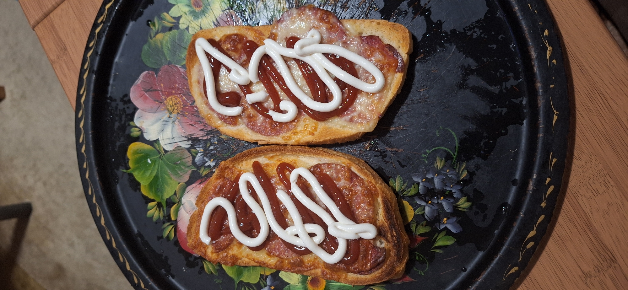 The image shows two open-faced sandwiches on a decorative black tray. The sandwiches appear to be made with sliced bread, topped with sliced sausage (possibly pepperoni), cheese, ketchup, and mayonnaise. The tray has a floral design with roses and other flowers in shades of pink, green, yellow, and purple. The tray is resting on a wooden surface. The image provides no information about the city of Blagoevgrad, Bulgaria.