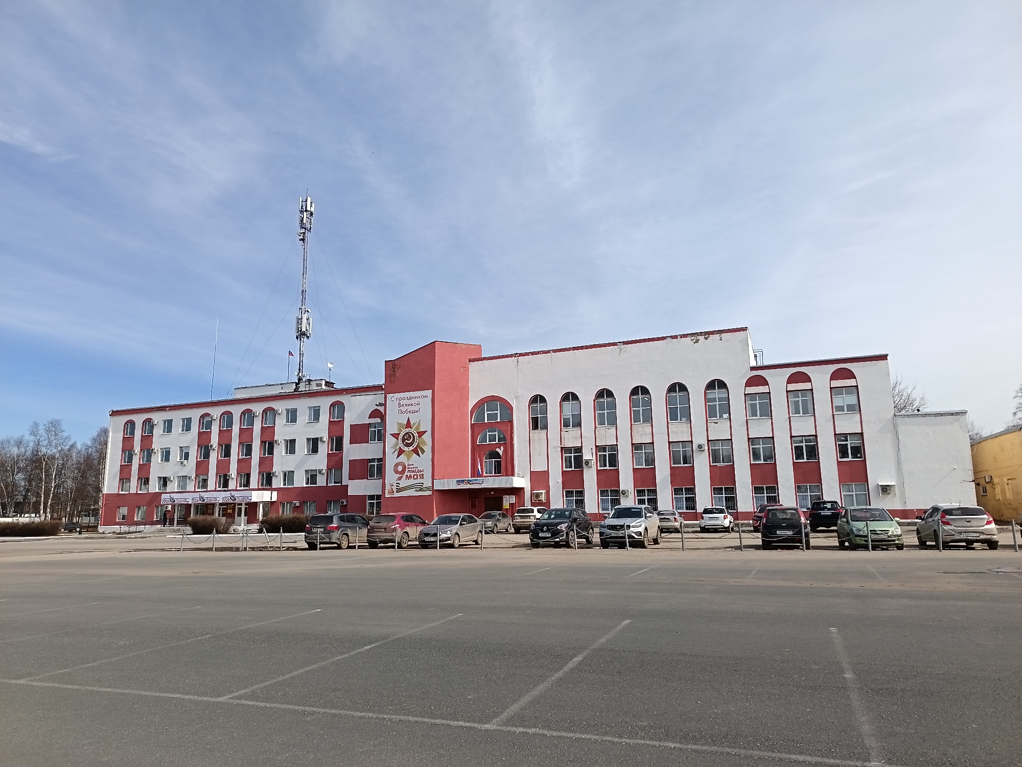 The image depicts a large, three-story building in Osa, Russia, characterized by its red and white striped facade. A communications tower stands to the left of the building. A wide, empty parking lot stretches out in front of the building, populated by several parked cars. A large sign adorns a central portion of the building, featuring a graphic of a red star, a hammer and sickle emblem, and Cyrillic text related to the celebration of "Victory Day" on May 9th. A row of trimmed bushes lines the base of the building. The sky above is blue with sparse clouds. No people are clearly visible.