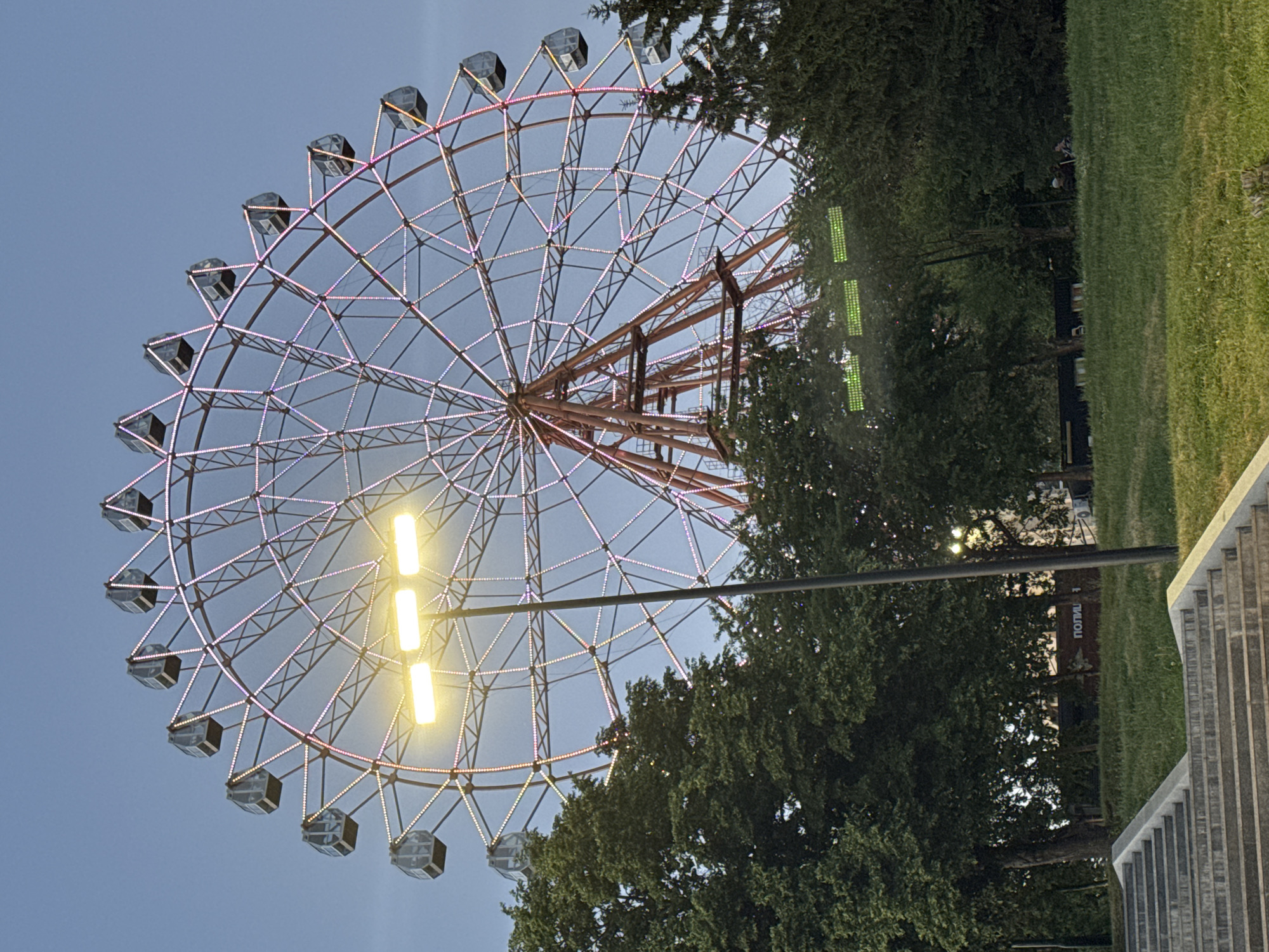 A large Ferris wheel is the central subject of this image, set against a clear blue sky. The wheel is adorned with numerous small lights, some of which are illuminated with pink and yellow hues, suggesting it is either dusk or nighttime. Several cabins, appearing dark and enclosed, are suspended from the rim of the wheel. The central structure of the Ferris wheel is a reddish-brown metal lattice.
To the right of the Ferris wheel, a dense cluster of dark green trees forms a backdrop, partially obscuring the wheel's structure. Beyond the trees, a strip of vibrant green grass can be seen, and further to the right, a section of a paved area with tiered steps is visible. There are also some bright green lights arranged vertically in the mid-ground, near the trees. A vertical pole runs through the image, intersecting the Ferris wheel.
In the background, obscured by trees, there appears to be a building or structure with some text visible on it. The visible text, appearing in white on a dark background, reads "ПОЛИЦ" which is the beginning of the word "Полиция" (Police) in Russian. This suggests the location might be near a police station or a public area with a police presence.
The overall scene appears to be an amusement park or a public park with recreational facilities. The time of day is likely evening, given the illumination of the Ferris wheel and the clear sky. No people are clearly visible in the frame.