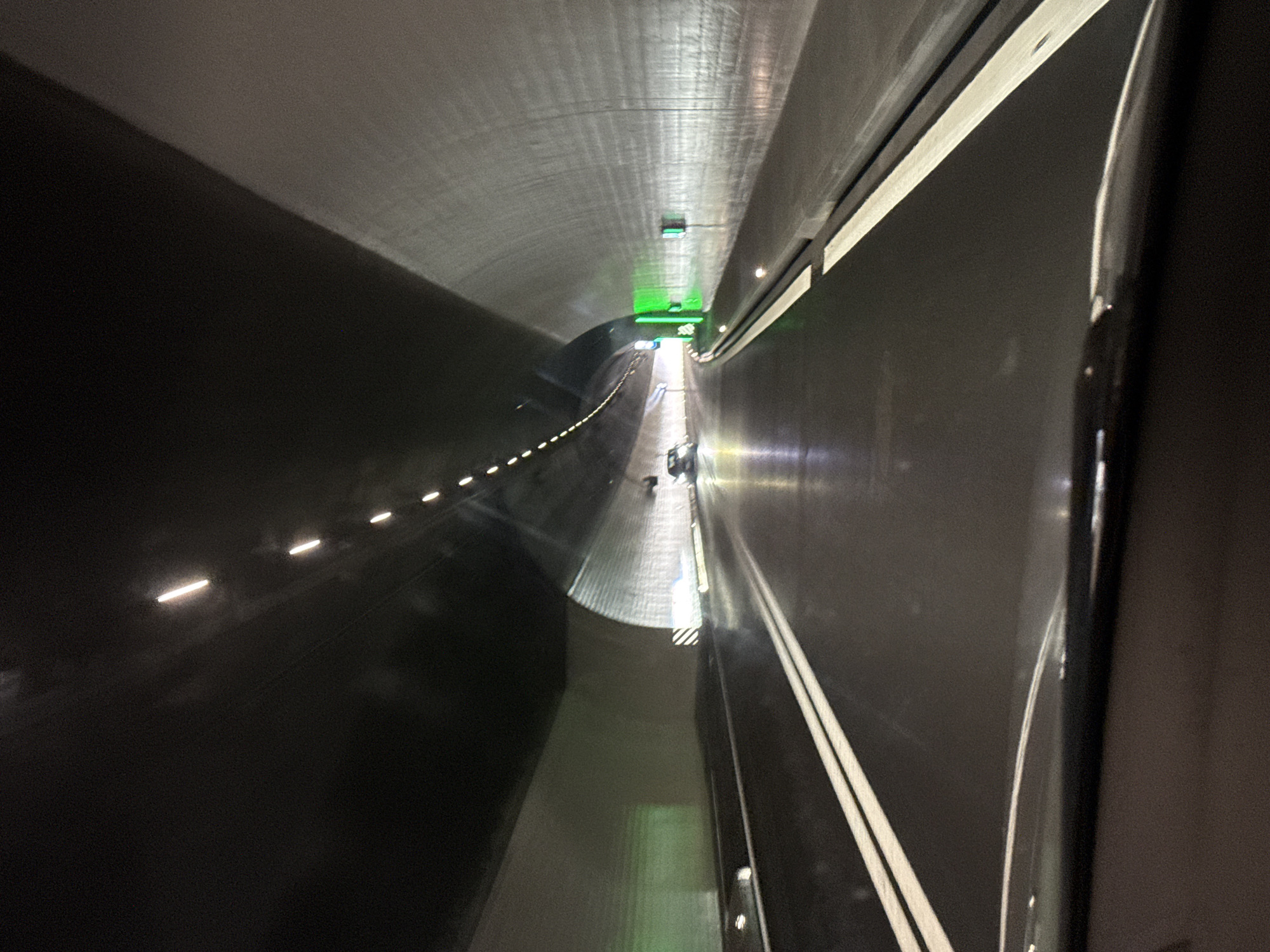 The image displays the interior of a long, concrete tunnel, captured from inside a moving vehicle with a slightly tilted perspective. The right edge of the frame reveals a dark, curved section of the vehicle's interior, likely a window frame.
The tunnel's walls and ceiling are a light gray, with striations visible, while the road surface is dark. Artificial illumination is prominent, featuring several green lights on the ceiling that project a strong glow, possibly indicating traffic signals or directional guidance. Along the left wall/roadway, a series of small, horizontally elongated white lights form a dashed line. On the right side of the dark road, two parallel white lines are painted, indicating a lane.
Further into the distance, at the end of the currently visible and well-lit section of the tunnel, two faint human figures can be seen standing or walking. A small, dark object, possibly a barrier or equipment, is also visible near them. Close to the center-right foreground, reflective white and black striped markings are on the road surface or a barrier.
The lighting within the tunnel is entirely artificial, contrasting with what appears to be daylight at the distant tunnel exit. The scene suggests travel through a modern, engineered pathway. The location context is Zurzach, Switzerland. There is no visible text in the image.
