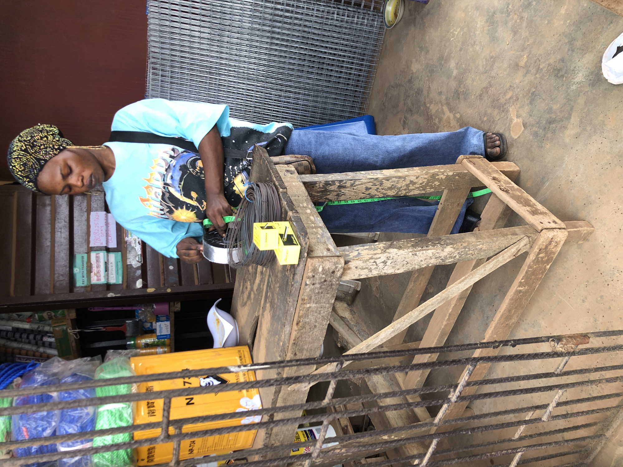 In Ijoko, Nigeria, a woman wearing a blue t-shirt and blue jeans stands behind a wooden table. She is holding wire and appears to be working with it. A roll of wire sits on the table, along with two small yellow boxes. Behind the woman, there is a wall of metal mesh. The floor is a dirt-colored concrete. The scene appears to be an outdoor market stall.
