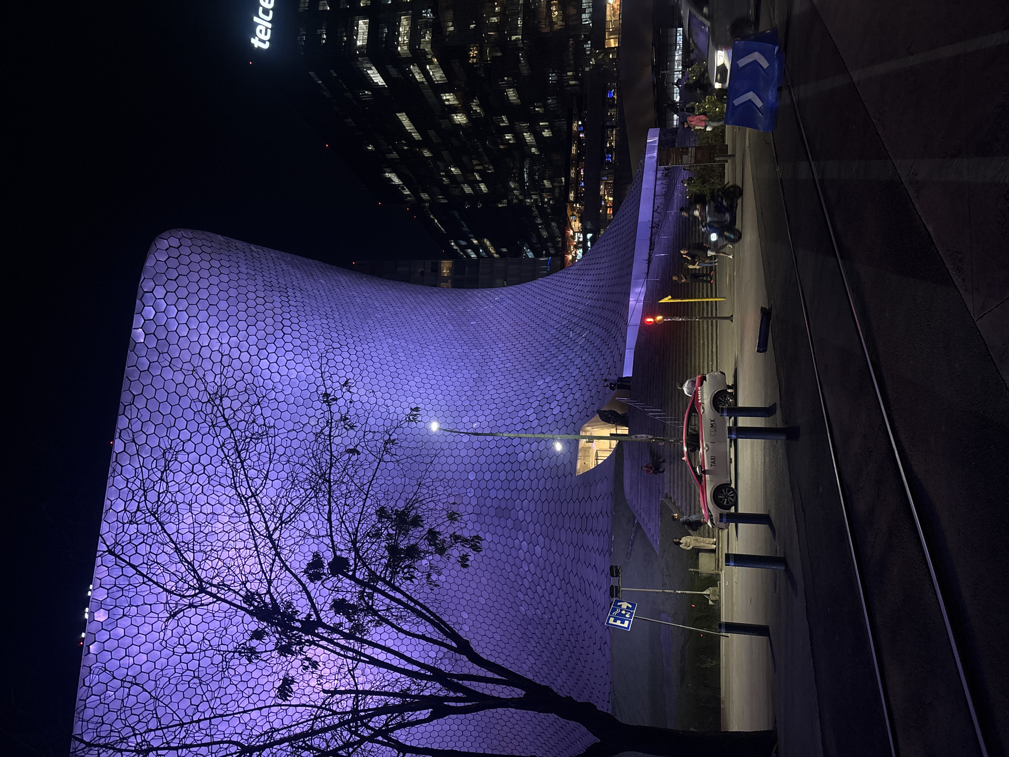 The image shows the Soumaya Museum in Mexico City, Mexico at night. The museum's distinctive, organically shaped building with a façade covered in hexagonal aluminum tiles is lit with a purple hue. In the foreground, bare branches of a tree frame the structure. A modern high-rise building, illuminated with many windows, is visible in the background, with the word "Telcel" displayed prominently. Along the right side of the frame, there is a road with a taxi parked on a curb. Several people are walking nearby. A traffic light is present and shows a red light. Street signs are visible.