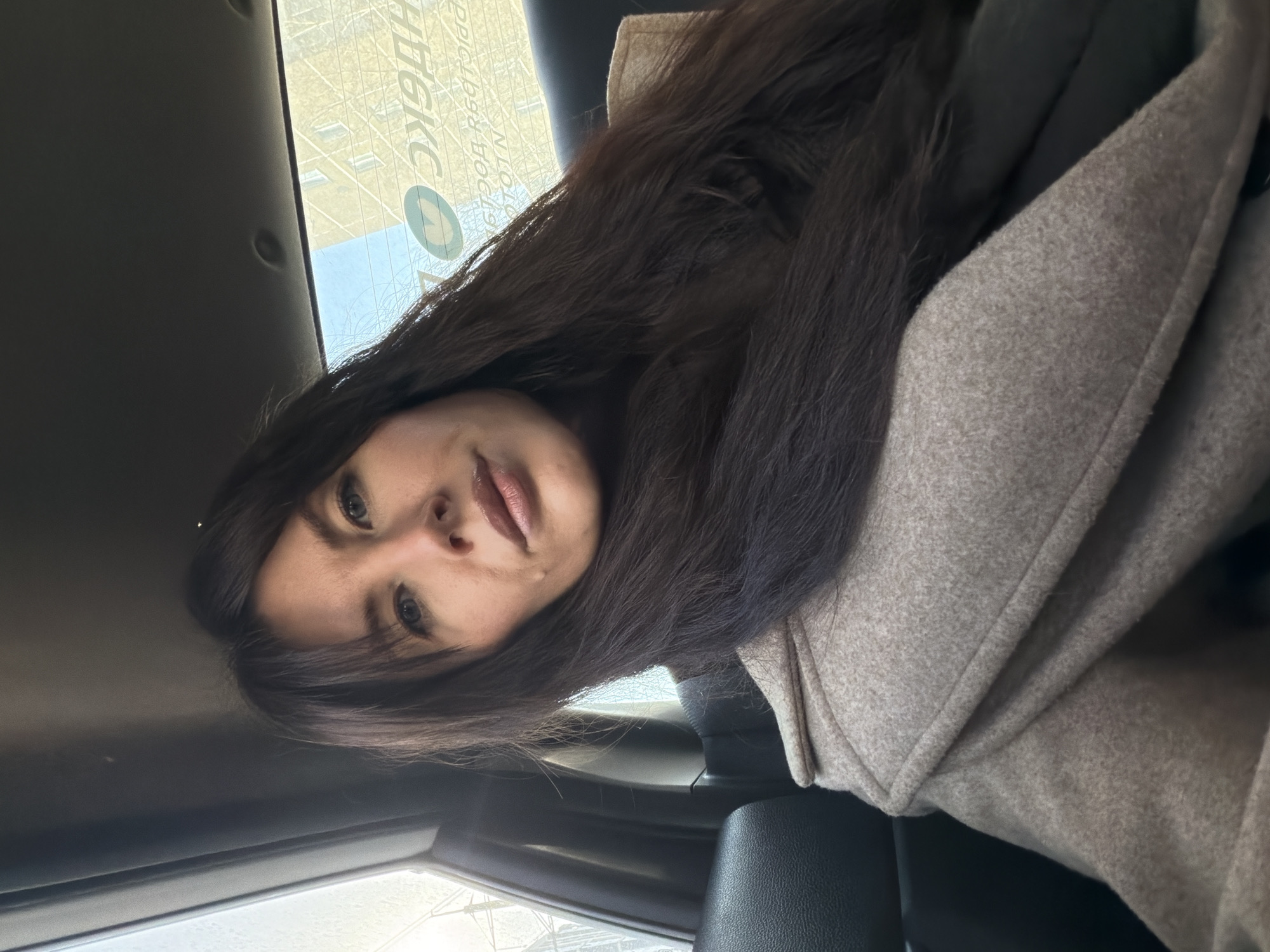 A female subject with long, dark brown hair and light-colored eyes is depicted taking a selfie in the backseat of a vehicle. She wears a light beige or grey wool-blend coat over dark clothing. The interior of the car features dark seating and a dark headliner. Through the rear window, a multi-story residential building with a light facade is visible under a clear sky. Reflective Cyrillic text and a circular logo, suggestive of a Yandex ride-sharing or delivery service, are present on the inside of the rear window, including partially legible reversed text like "ДОСТАВКА" (Delivery) and "ОТОПЛЕНИЕ" (Heating). The vehicle is situated in the city of Perm, Russia.
