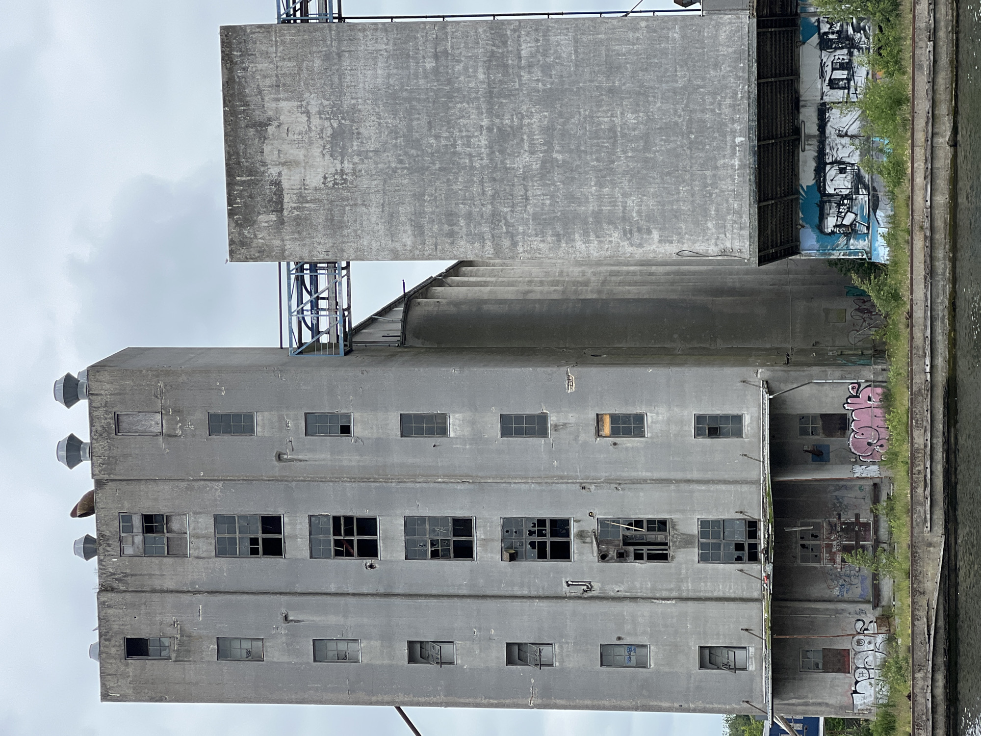 This is a medium shot of the abandoned Nordværket industrial complex in Odense, Denmark. The main structure is a tall, multi-story concrete building with many broken windows and signs of decay. Adjacent to it is a wider, shorter concrete silo with a textured surface. A metal walkway connects the two structures. The base of the buildings is covered in graffiti, and overgrown vegetation grows in front of them. A body of water is visible in the foreground. The sky is overcast. There are no people or animals visible in the image.