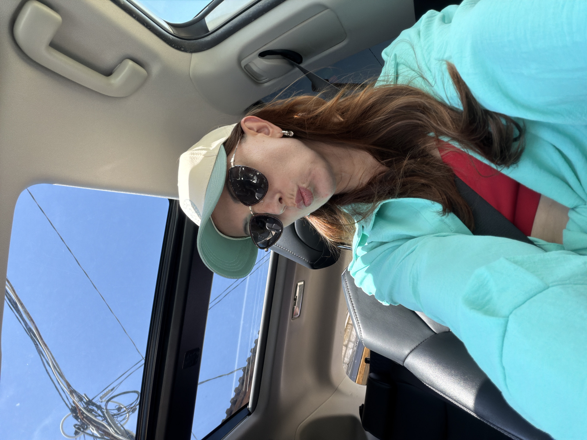 A woman is taking a selfie inside a car with a sunroof. She is wearing sunglasses and a baseball cap. She has shoulder-length brown hair. She is wearing a light blue shirt. The car has black leather seats. Outside the sunroof, there are power lines against a blue sky. There is no evidence to indicate this image was taken in Birobidzhan, Russia.