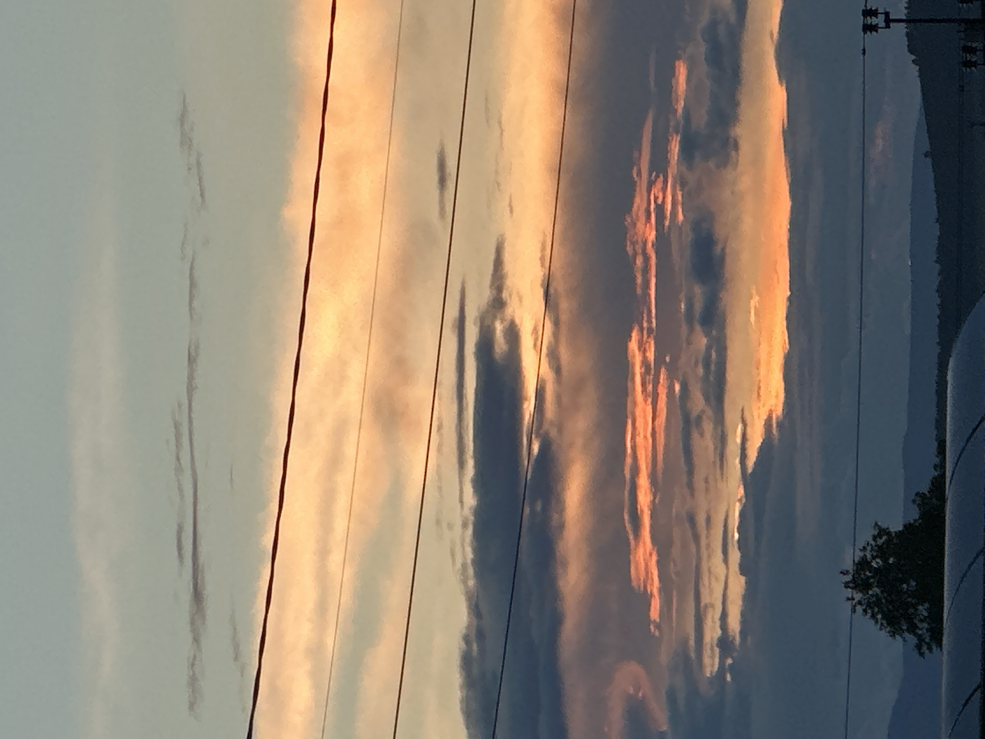 The image depicts an outdoor scene at either dawn or dusk, located in the city of Unknown, Unknown. The sky is filled with horizontal cloud formations, transitioning from a light blue-grey in the upper section to vibrant orange and pink hues interspersed with dark grey clouds in the middle and lower portions. Multiple dark, thin wires, likely power lines or communication cables, stretch horizontally across the frame at various elevations. In the lower left quadrant, a dark, silhouetted tree is partially visible above a curved, dark structure, possibly a roof. On the far right edge, a section of a utility pole with attached insulators and crossarms is present. The background reveals a silhouette of distant hills or mountains along the horizon. No people or discernible activities are present.