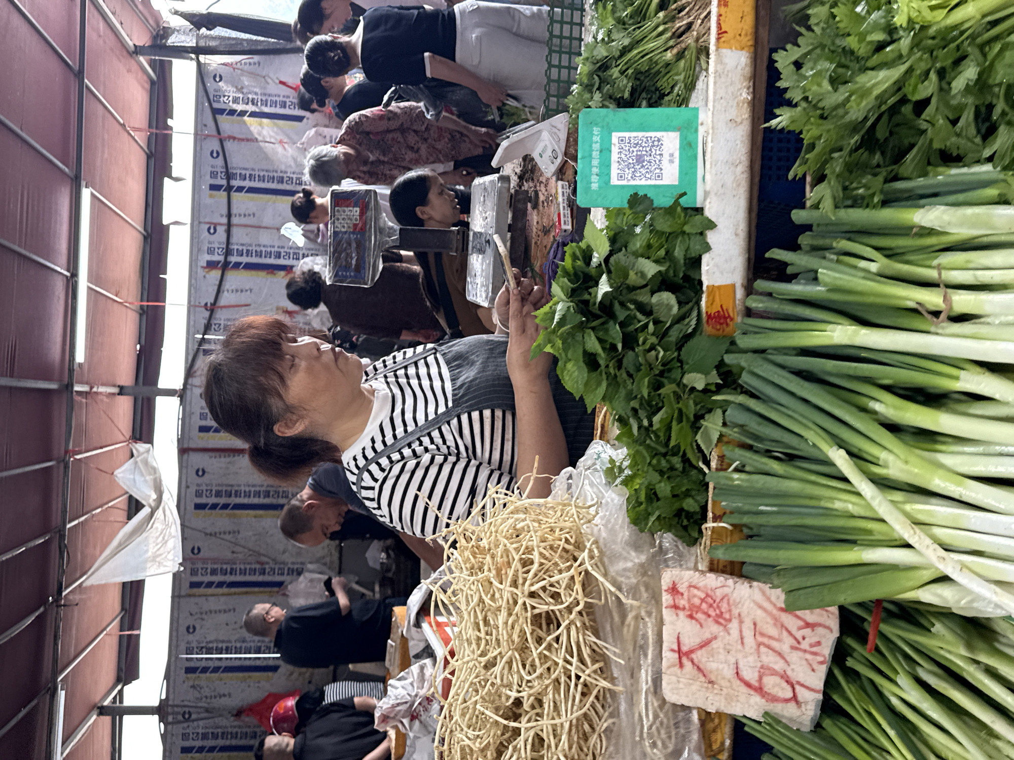 This image captures a bustling outdoor market scene, likely in Jiangjin, China. A woman with dark hair pulled back into a ponytail, wearing a striped shirt and an apron, is the central figure, engaged in weighing produce on a scale. Surrounding her are various fresh vegetables, including bundles of long, thin roots, leafy greens, and bunches of green onions. A sign with Chinese characters and a QR code is visible, suggesting a modern payment option. In the background, other shoppers and vendors are present, with a backdrop of market stalls and what appear to be printed banners. The overall environment suggests a typical day at a local food market, with an emphasis on fresh produce and commerce.