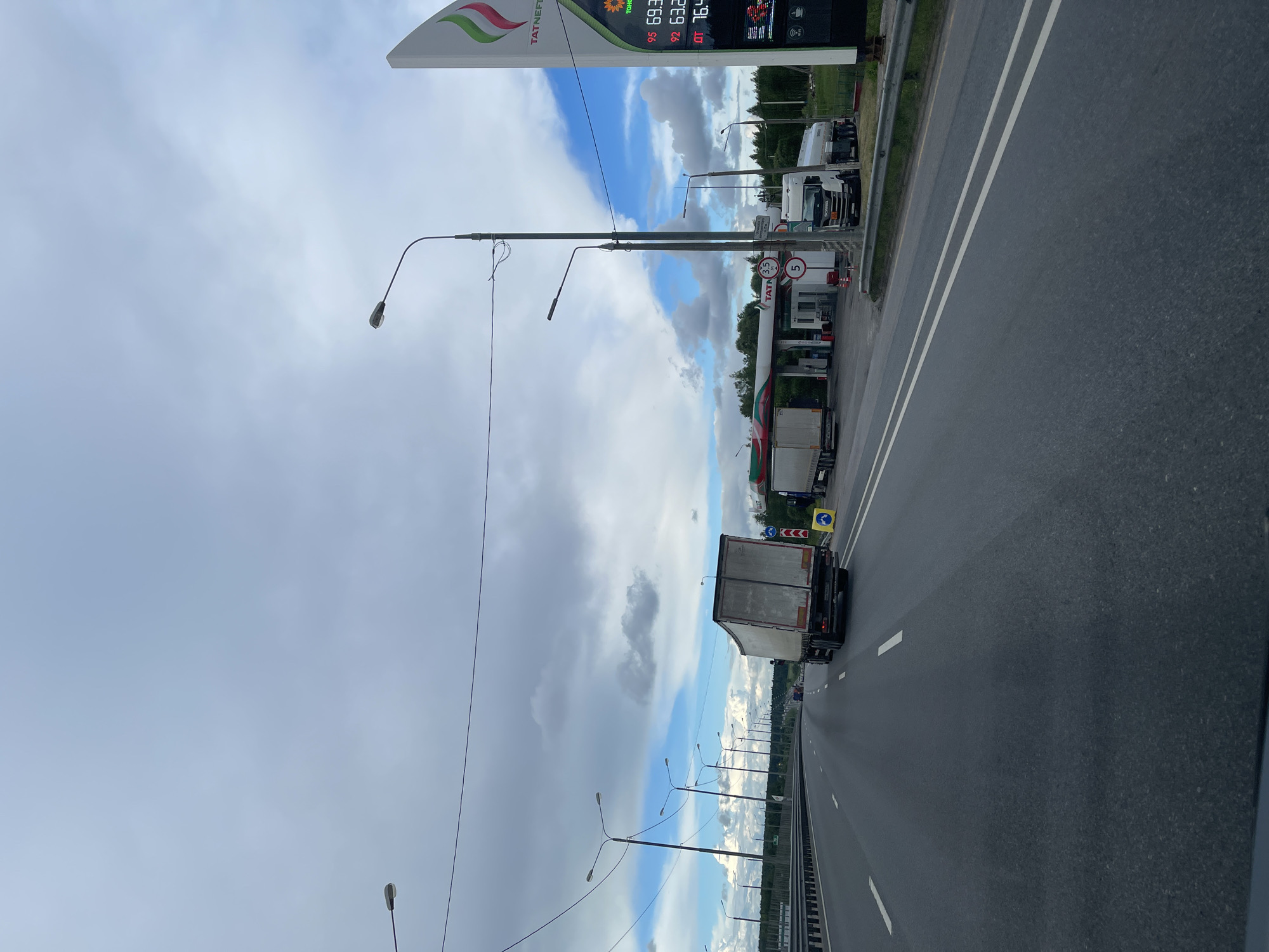The image depicts a highway in the vicinity of a Tatneft gas station in Бавыкино, Russia. A white truck is traveling on the rightmost lane of the highway. Street lights line the road's edge. The sky is cloudy. The gas station displays fuel prices on a sign. Several other trucks are parked at the gas station.