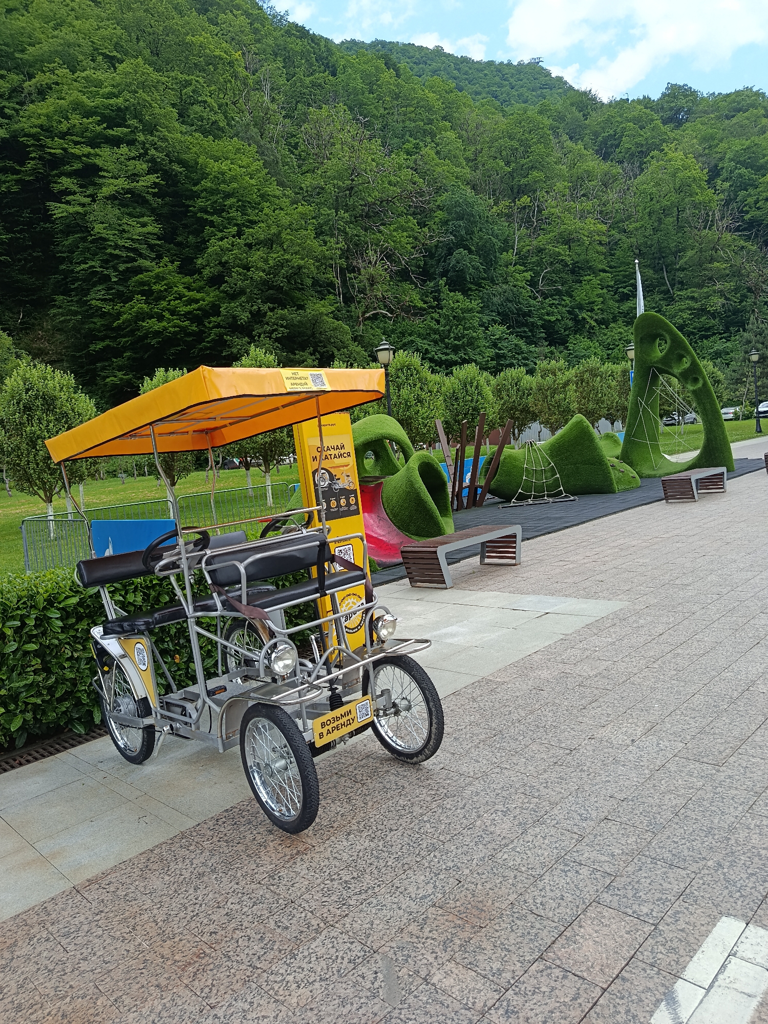 The image shows a family-sized peddle cart with a yellow canopy parked on a paved walkway in the city of Estosadok, Russia. The peddle cart is in the foreground, with a dense green forest covering a hillside in the background. To the right of the cart, there is a playground with unique, sculpted green structures that resemble hills and tunnels, along with benches and a net for climbing. A red slide is also visible as part of the playground. The sky is partly cloudy. The overall scene depicts a recreational area in a mountainous, forested environment.