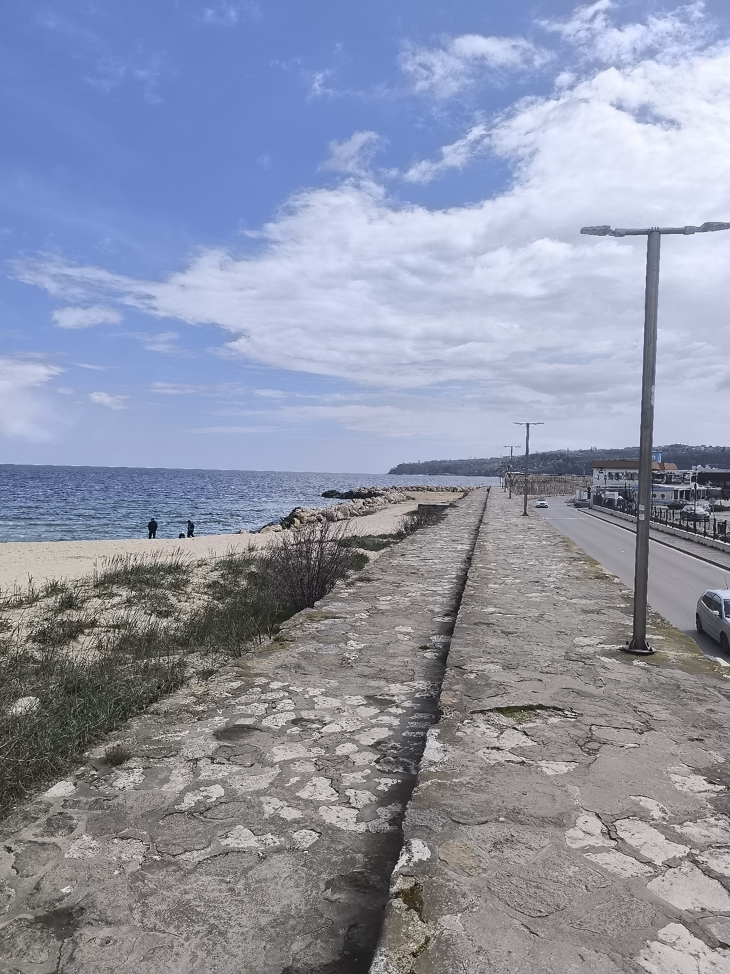 This image depicts a coastal scene in Varna, Bulgaria. The perspective is from a raised stone pathway or sea wall. To the left of the pathway is a sandy beach with dune grass and a few small shrubs. Two individuals are standing on the beach near the water's edge, possibly fishing. The sea extends to the horizon under a sky with scattered clouds. To the right of the pathway is a street with several street lights and some parked and moving vehicles. A line of buildings and trees are visible in the distance along the coastline. The stone pathway is uneven and appears aged.