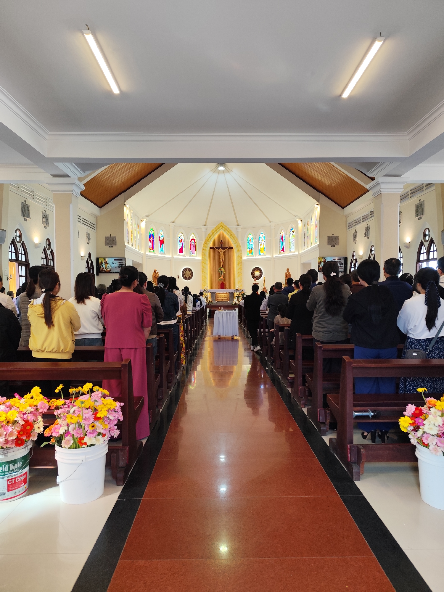 Inside a church in Lâm Viên - Đà Lạt, Vietnam, congregants stand in rows of wooden pews during a service. The floor is a glossy brown tile, bordered by white tiles. Two buckets of colorful flowers are placed on either side of the aisle at the forefront. The church features stained glass windows and a large golden crucifix at the altar. Overhead, fluorescent lights illuminate the interior.