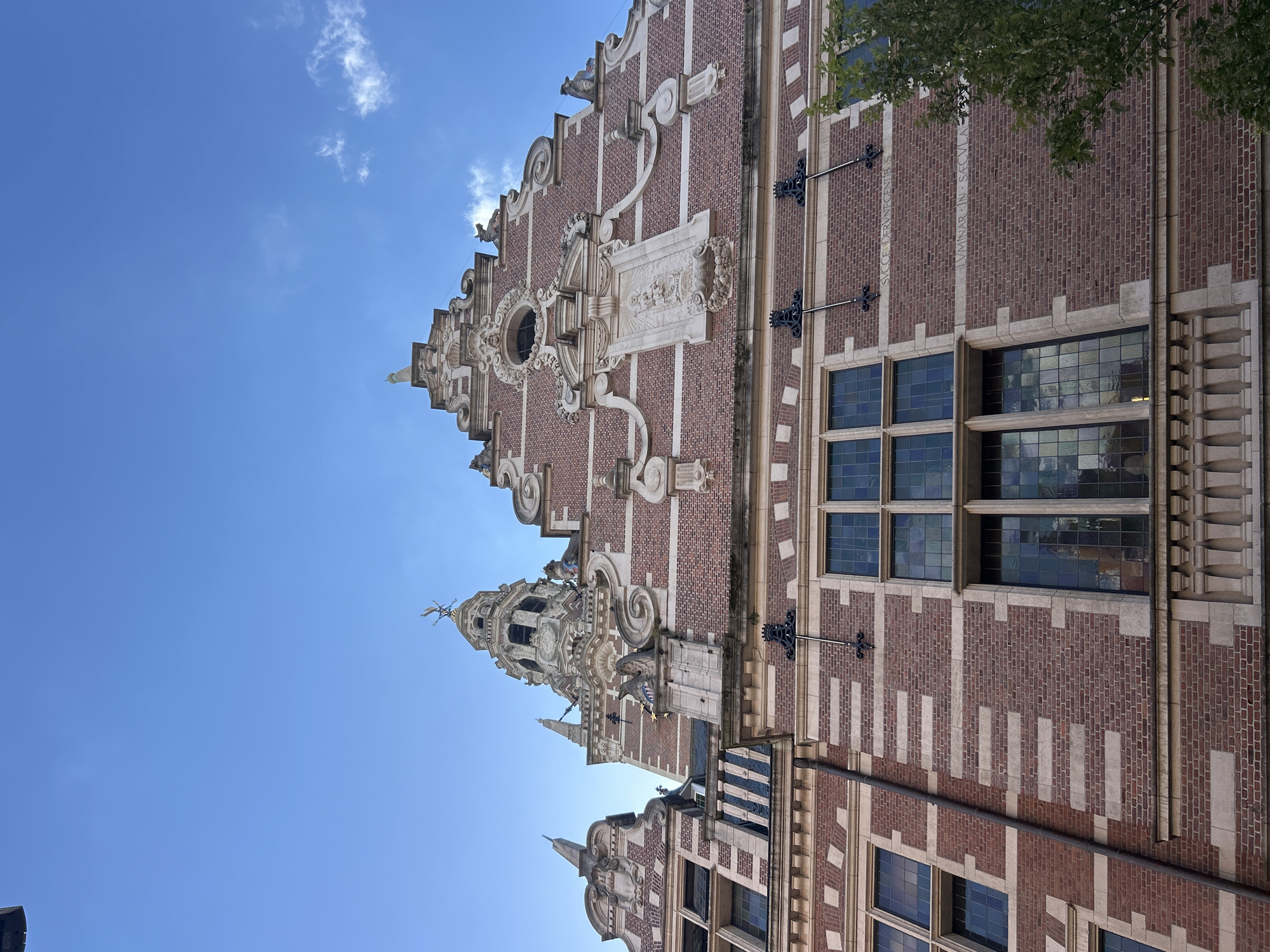 The image shows the exterior of the University Halls building in Leuven, Belgium, viewed from below against a clear blue sky with a few wispy clouds. The architecture is characterized by red brickwork with lighter stone accents and ornate decorative elements.
The building features stepped gables, prominent arched windows with multiple panes, and a tall, multi-tiered tower with a spire and weather vane. Sculptural details, including what appear to be statues of figures and mythical creatures, are visible on the facade. Inscriptions in Latin are present on the brickwork. A pipe runs vertically down the side of the building, and a tree with green foliage is partially visible on the right side of the frame. The perspective emphasizes the grandeur and historical nature of the building.
