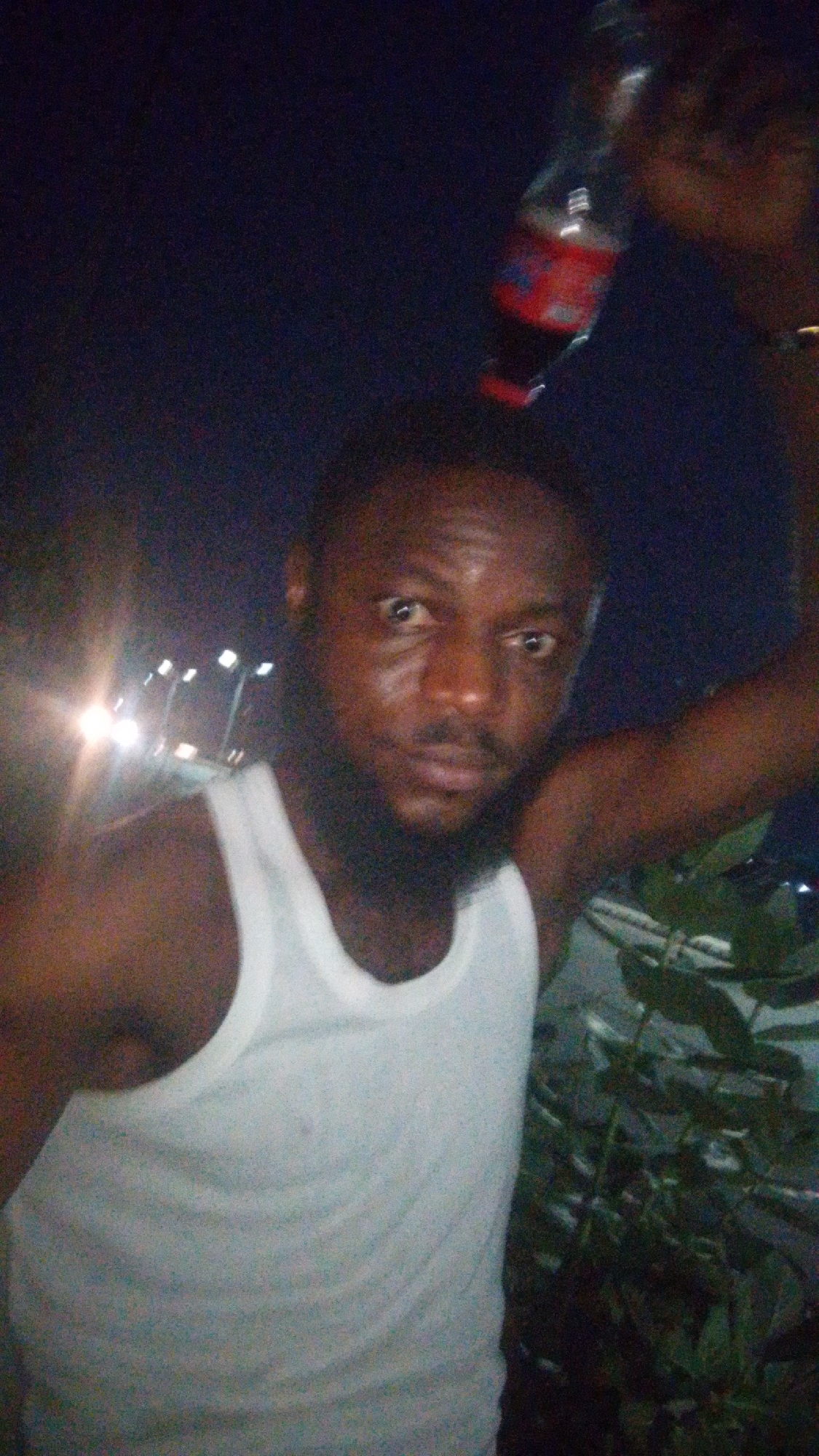 A photograph taken at night in Lokoja, Nigeria, shows a man in a white tank top, a dark beard, and short hair facing the camera. He is holding a bottle of Coca-Cola above his head. Behind him and to the left are streetlights. There are green plants near the bottom right of the image. Another person's face is slightly visible in the upper right of the frame near the bottle.