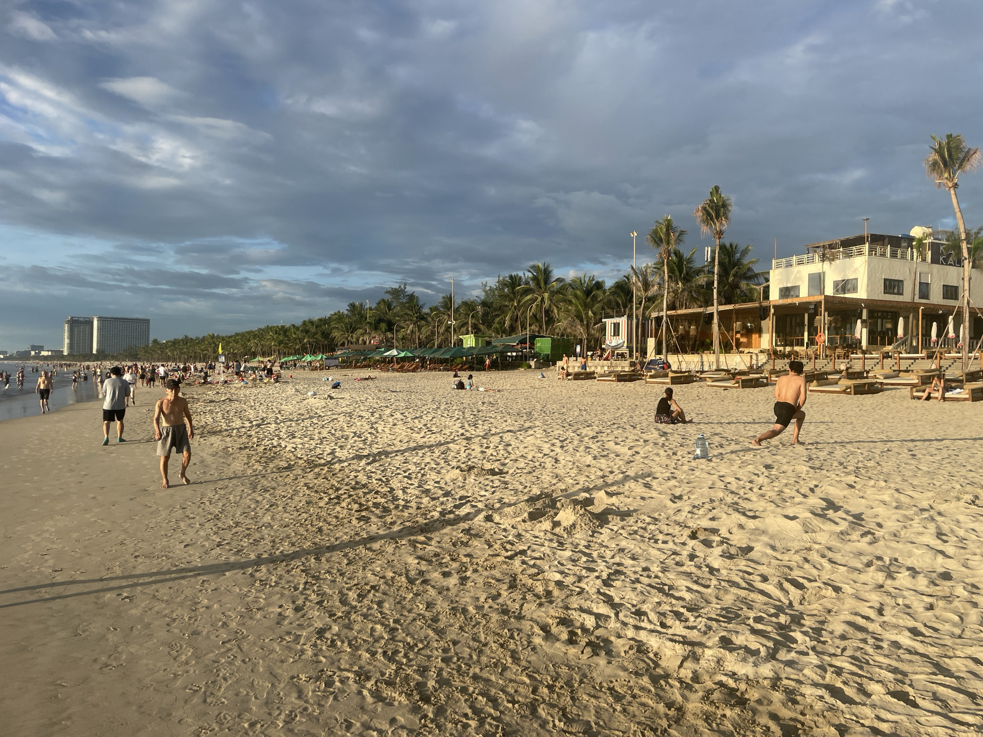 This image depicts a beach scene in Đà Nẵng, Vietnam. The sandy beach is populated with numerous people enjoying the day, some walking, others relaxing or engaged in recreational activities. In the background, a line of palm trees stretches along the coastline, with a large, modern hotel or resort visible in the distance to the left. To the right, a multi-story beachfront establishment with an open-air ground floor and a white upper level, featuring balconies and awnings, is prominent. Beach umbrellas and lounge chairs are set up on the sand in front of this building and further down the beach. The sky is partly cloudy with a mix of blue sky and dark, dramatic clouds, suggesting either early morning or late afternoon light. The tide appears to be out, revealing a wide expanse of sand. Several individuals are captured in motion, such as two men playing what appears to be a ball game in the mid-ground.
