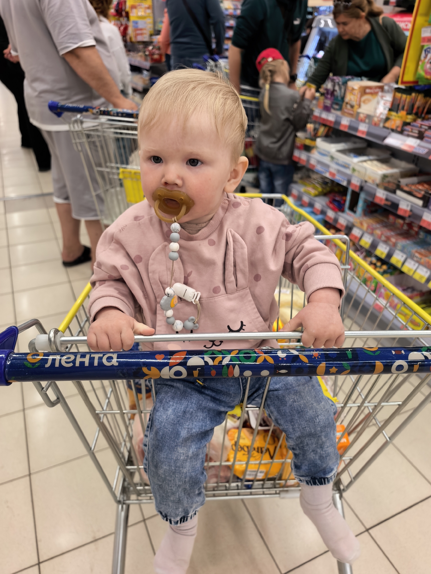 A close-up, eye-level photograph captures a blonde-haired infant or toddler seated in the child's seat of a supermarket shopping cart. The child wears a pink long-sleeved sweatshirt with dark pink polka dots and what appear to be bunny ear details on the hood, blue denim jeans, and light pink socks. A brown pacifier is held in the baby's mouth, secured by a decorative clip with grey and white beads and a small animal-shaped charm, visible against the pink sweatshirt. The child grips the blue plastic handle of the shopping cart with both hands, looking towards the left side of the frame with a neutral, slightly contemplative expression.
The blue cart handle features colorful, whimsical patterns and the prominent white Cyrillic text "ЛЕНТА" (LENTA), identifying the store as a Lenta hypermarket. The background is softly blurred but reveals typical supermarket aisles with shelves stocked with various consumer products. Several other shoppers, including adults and another child, are visible in the distance, browsing or moving through the store. The lighting suggests an indoor, daytime setting. The scene depicts a common grocery shopping activity in Sterlitamak, Russia, with the baby as the focal point within the retail environment.
