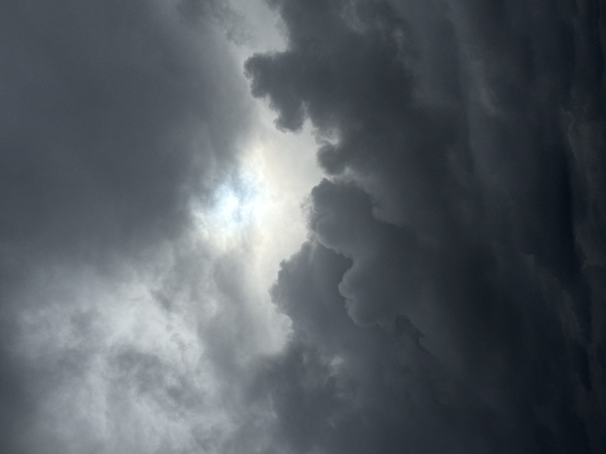 The image displays a sky filled with a dense formation of dark grey and cumuliform clouds, indicative of an overcast or impending storm condition. A distinct, irregular patch of brighter, lighter-grey sky, possibly revealing sunlight, is visible in the upper-center quadrant, partially obscured by the surrounding darker cloud masses. The lower portion of the sky appears significantly darker and more uniform in its cloud cover. No land features, structures, people, or objects are visible within the frame. The scene is situated over the city of 's-Gravenzande, Netherlands.