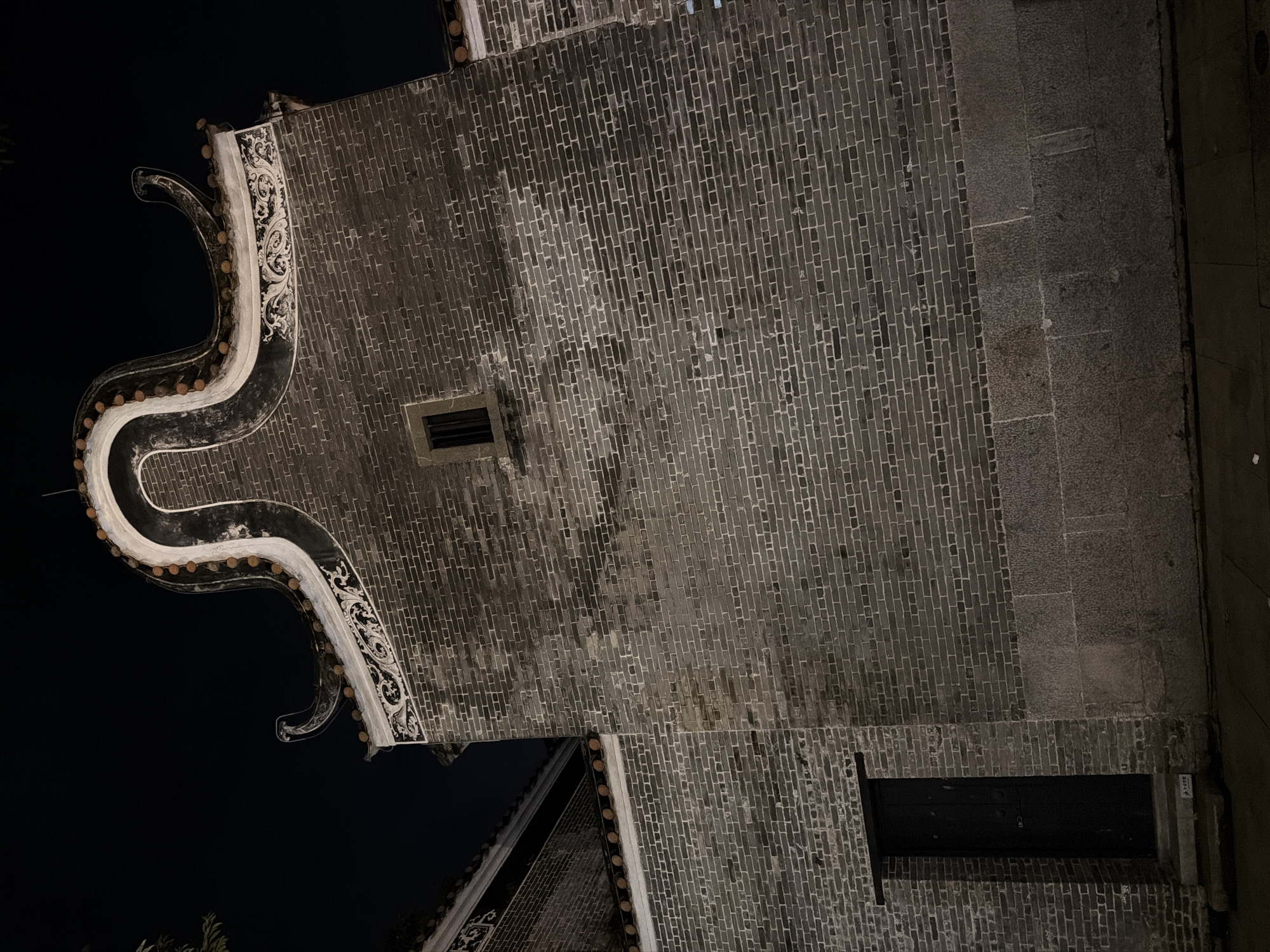 A low-angle, nighttime, full-shot photograph captures the facade of a traditional Chinese building in Chancheng District, China. The building is constructed primarily from dark grey bricks, with a stone base. A black wooden door is visible on the left side of the image, set within a brick wall, and is approached by a few steps. A small, barred window is positioned higher up on the brick wall. The roofline features a decorative, curved gable adorned with white plasterwork and small orange tiles. The overall scene is illuminated by artificial light, casting shadows and highlighting the textures of the materials. The sky is dark and unlit.