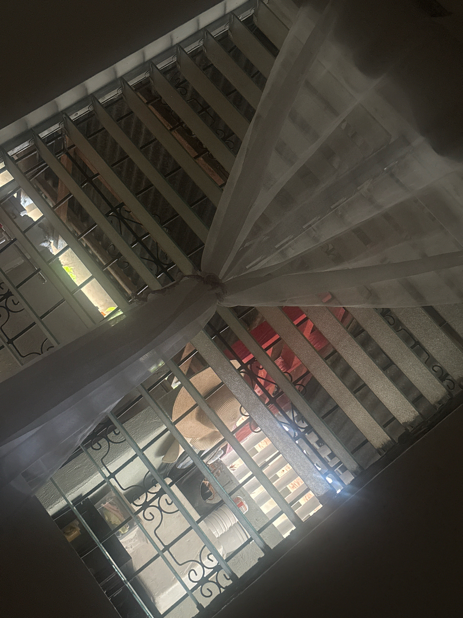 The image captures an upward view of a window grate covered by a sheer white fabric, possibly a mosquito net, creating a draped effect. Through the grate and fabric, various objects are visible, including what appears to be a straw hat, a red object, and what could be clothing or fabric. The lighting suggests it is daytime, with light filtering through the window. The setting is identified as being in Santa Cruz, Philippines, hinting at a residential or indoor location. The angle of the shot is from below, looking up towards the window.
