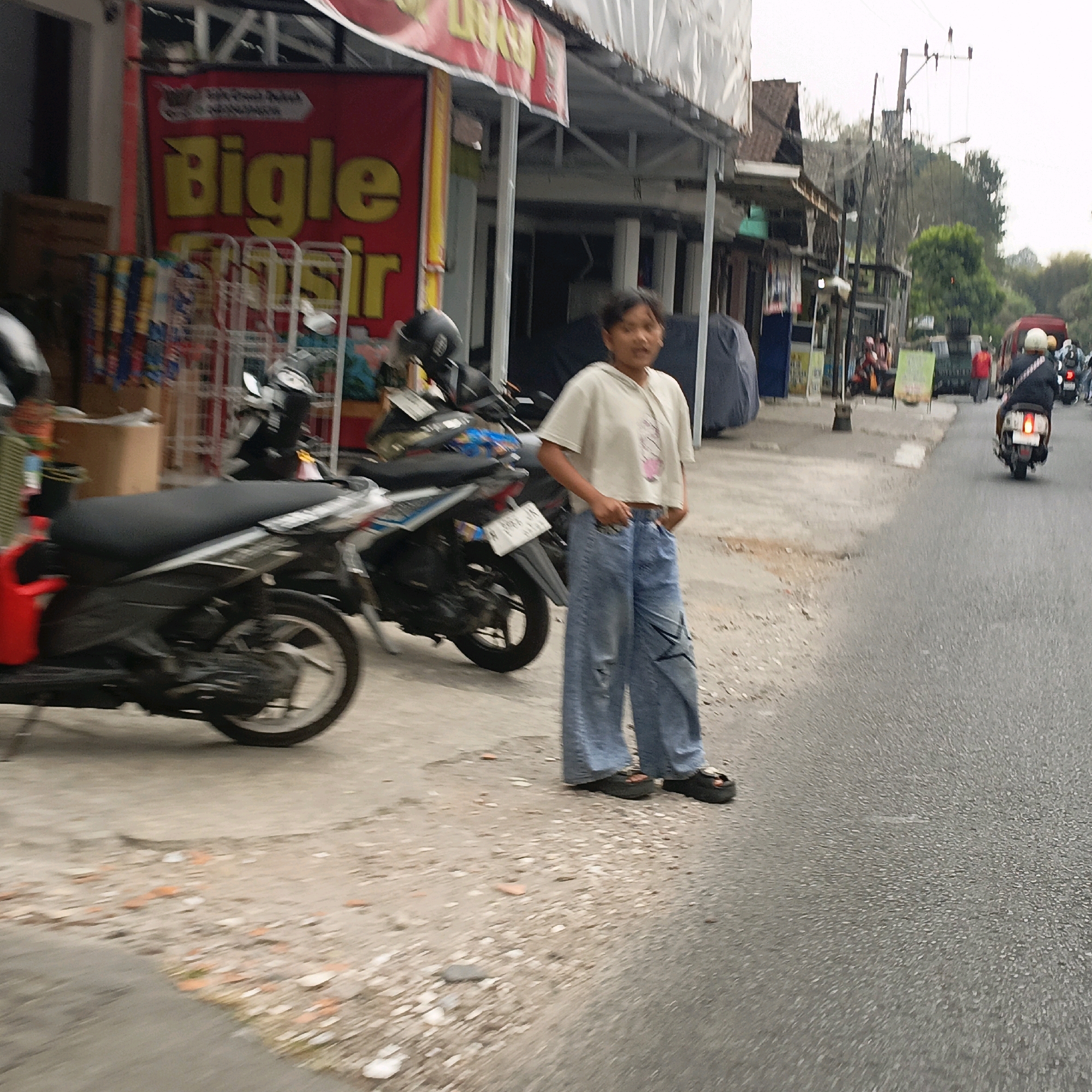 A young person stands on the side of a street in Salatiga, Indonesia, looking to their right. They are wearing a cropped, light-colored hooded sweatshirt with a graphic on the front and loose-fitting, distressed denim jeans. The person is also wearing platform sandals. To their left, there are parked motorcycles. In the background, a red banner with yellow text that reads "Bigle Sir" is visible, along with storefronts. The street itself appears to be paved, with some debris and gravel on the shoulder. Traffic, including other motorcycles and a van, can be seen further down the road. The overall scene depicts a typical street view in an Indonesian city.