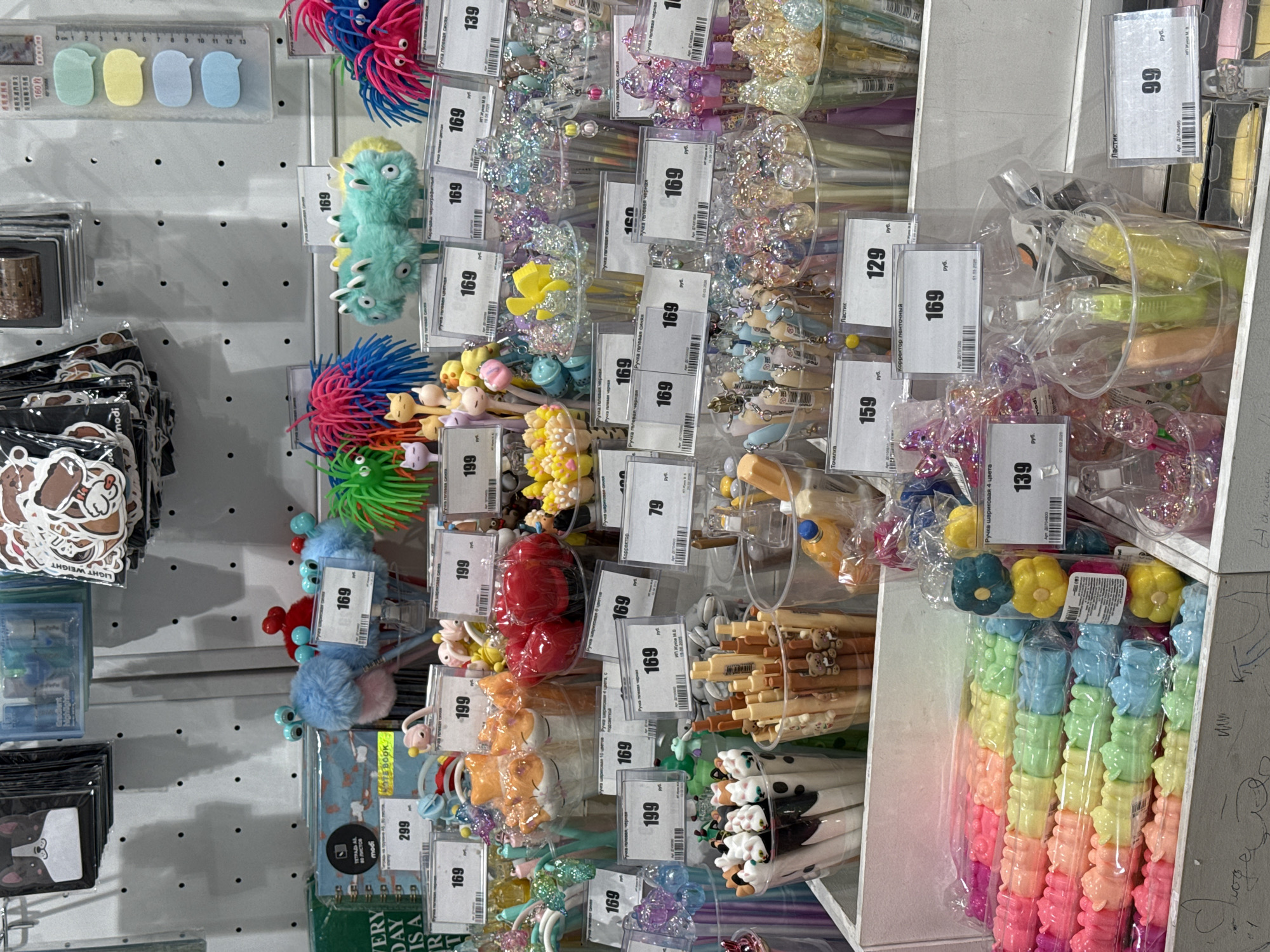 The image depicts a retail display in a store, likely located in Saint Petersburg, Russia, based on the Cyrillic text and currency symbol "руб" (rubles) on the price tags. The scene focuses on a variety of stationery items, primarily pens with decorative toppers in clear plastic containers. The pens feature colorful, whimsical designs, including cartoon characters, animals, flowers, and beads. Prices vary, with tags indicating costs such as 129, 139, 159, 169, 199, and 299 rubles. Other stationery items visible include stickers, erasers, and correction tape. The background is a white, perforated wall, commonly found in retail environments.