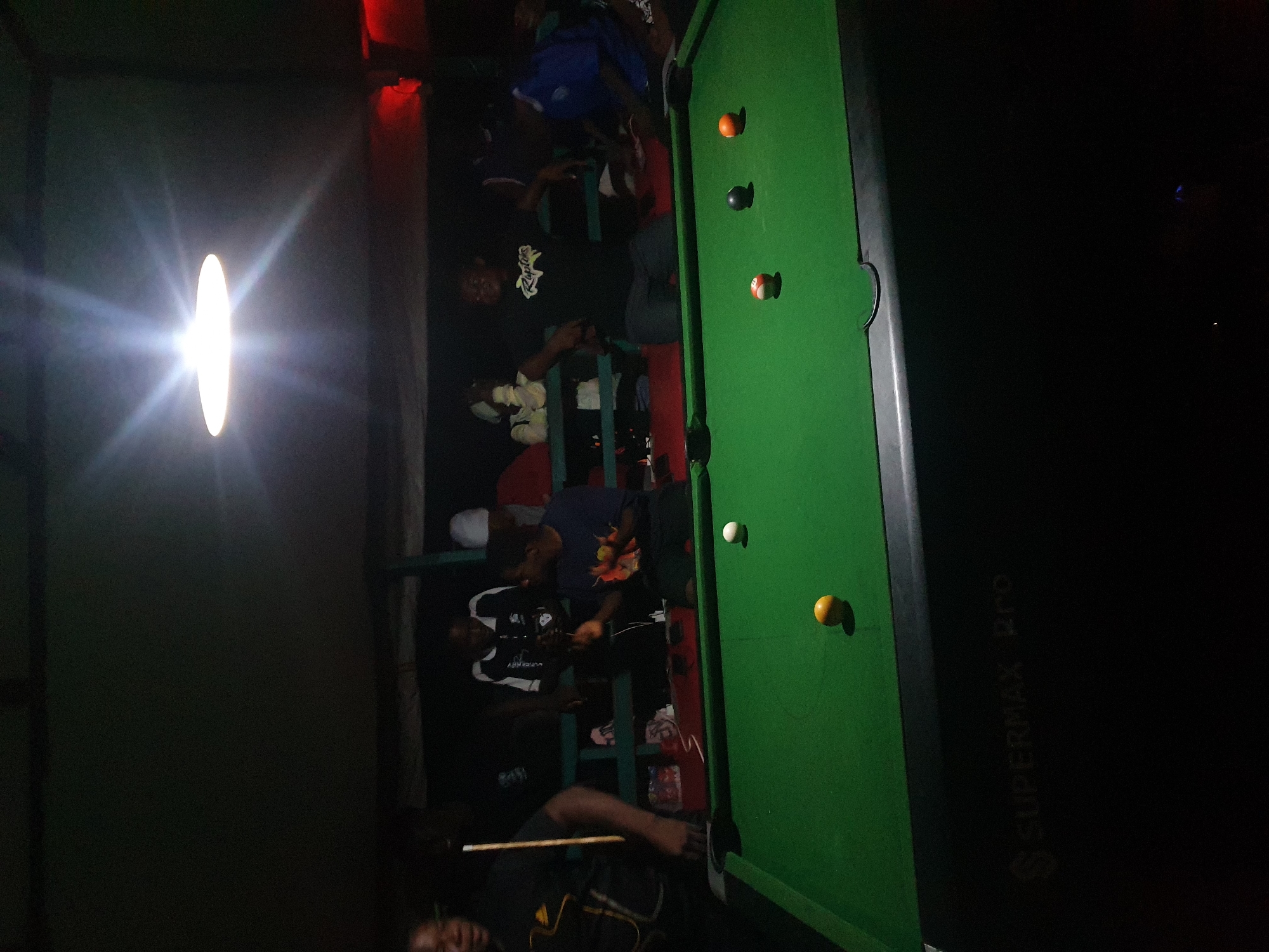 In Lokoja, Nigeria, this nighttime scene depicts a group of individuals gathered around a pool table inside a dimly lit establishment, possibly a bar or recreation center. A bright overhead light illuminates the pool table, which is covered in green felt and has several balls on it: a white cue ball, an orange ball, a black ball, a striped ball, and a yellow ball. Two of the balls are positioned near the center of the table, while others are scattered.
In the foreground, the edge of the pool table and the underside are visible, with the "SUPERMAX Pro" branding subtly displayed on the table's casing. Behind the table, several people are standing or casually leaning. One individual on the left, facing away from the camera, wears a black t-shirt and holds a pool cue. Another person in the center wears a black t-shirt with a graphic design and appears to be engaged in conversation. To their right, a person in a blue shirt is also visible. Other individuals are partially obscured in the background, with some appearing to be watching the game. The overall ambiance suggests a relaxed, social gathering with a focus on the game of pool.