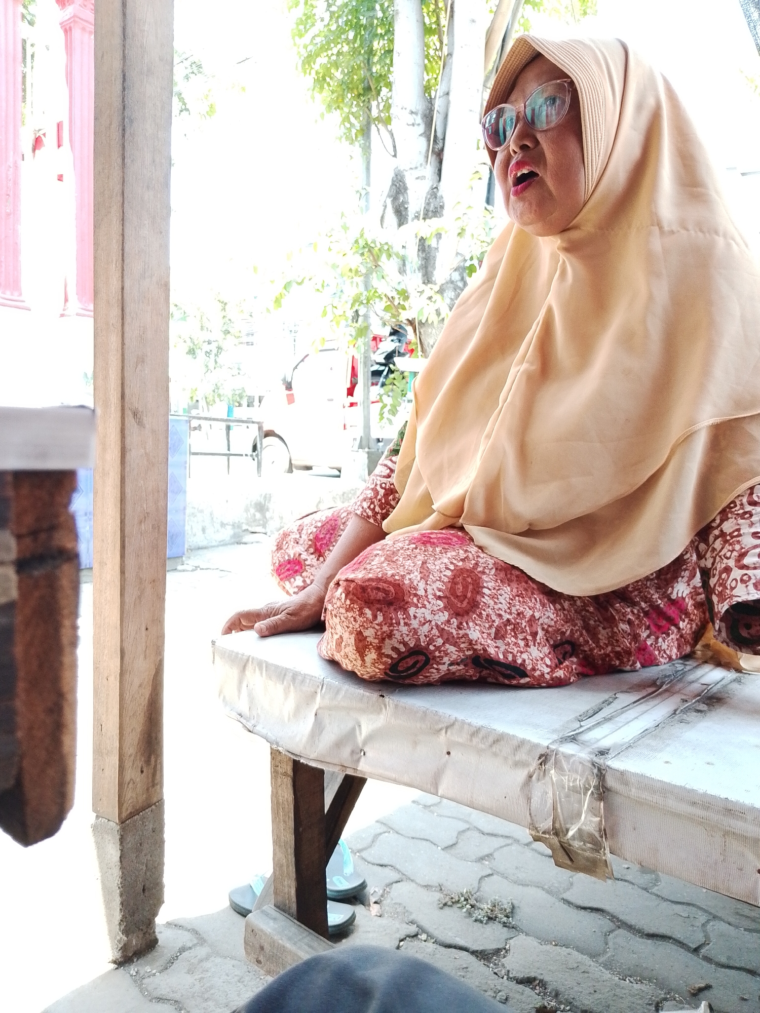 An outdoor medium shot captures an Indonesian woman, wearing glasses and a light tan hijab, seated on a white table in what appears to be an outdoor market in Pasuruan, Indonesia. Her mouth is open, and her head is turned to her left, suggesting she is speaking. She wears a red and white patterned dress and her right hand rests on her thigh. The table is covered in a white material that is taped down with brown tape. Behind the woman, trees and a pink building are visible. A wooden post is to the left of the frame, and a pair of light blue flip-flops are on the ground below the table. The ground is paved with gray interlocking bricks.
