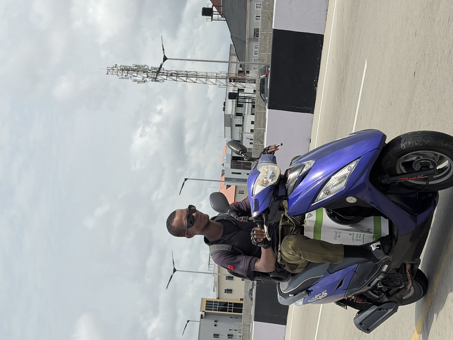 The image captures a young man sitting on a blue scooter in an outdoor urban setting in Alaguntan, Nigeria. The photograph is oriented vertically, showing the man and scooter tilted 90 degrees to the right.
The central subject is a man with dark skin and a short haircut, wearing dark sunglasses that obscure his eyes. He is dressed in a dark short-sleeved top or jacket, fingerless gloves, and olive-green cargo-style pants. He sits upright on the scooter, holding the handlebars with both hands, suggesting he is either paused or posing.
The scooter itself is a vibrant metallic blue or purple, with "Disk" visible on the front wheel and "KRS" on the side panel. A white and green bag, possibly a delivery or shopping bag, is placed between the man's legs on the scooter's floorboard. The bag features the word "Flourish" prominently, along with a partially visible phone number starting with "+234 81..." and other smaller text and logos.
The background reveals a slightly overcast or cloudy sky, indicating daytime. The ground is a light-colored concrete surface with white painted lines, suggestive of a parking lot or a wide paved area. In the distance, there are various buildings, including multi-story residential or commercial structures, and a prominent tall metal communications or cell tower. Several streetlights, some appearing to be solar-powered, are also visible. A low wall, painted black and white, separates the immediate foreground from the buildings further back. The overall impression is a bustling yet currently calm urban environment.