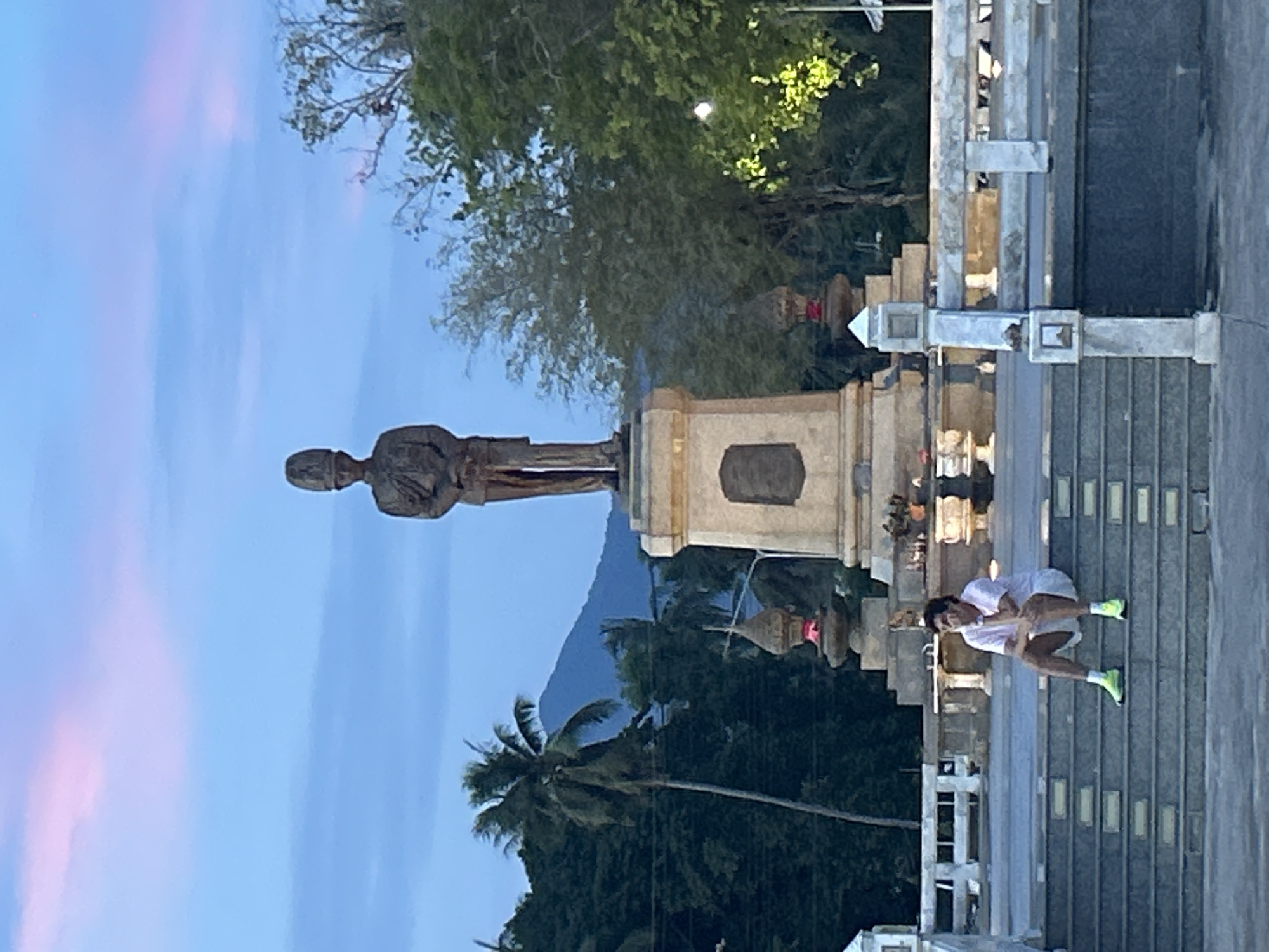 A statue stands on a multi-tiered pedestal in Thong Sala, Thailand. A person is kneeling on the steps of the structure. Trees and a mountain are in the background with a dusky sky overhead. The statue is of a person in uniform. The kneeling person is wearing white clothing and green shoes. The structure's steps are gray.