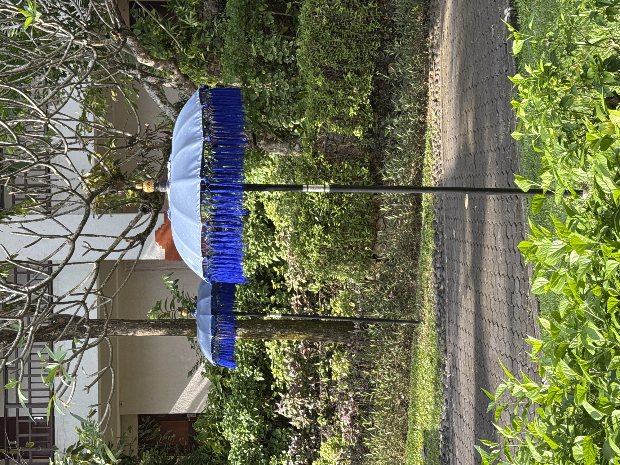 The image shows two light blue Balinese parasols with dark blue fringe standing in an outdoor location near Kuta Selatan, Indonesia. The parasols are situated beside a paved walkway with grassy areas on either side, surrounded by lush tropical greenery including trees and various shrubs. A building with doors and windows is visible in the background.