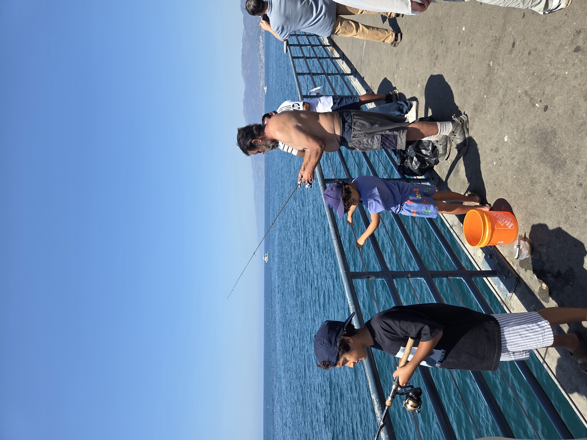The image depicts a sunny day on a pier in Santa Monica, California. Several people are present, primarily engaged in fishing.
In the foreground, a young boy wearing a dark t-shirt and striped shorts stands with a fishing rod. To his right, a younger child in a blue t-shirt and patterned shorts is also fishing, with an orange bucket nearby.
Further back, a shirtless man with a beard, wearing shorts, is fishing. Behind him, another person in a white shirt and shorts is visible. To the far right, a man in a blue shirt and khaki pants holds a phone up, seemingly taking a picture, and another person in a light-colored outfit is partially visible.
The pier is made of concrete, with a metal railing separating the walkway from the ocean. The water is a deep blue, with a few whitecaps. In the distance, mountains are visible under a clear blue sky. A small white boat is present on the water. The overall scene suggests a casual recreational activity.