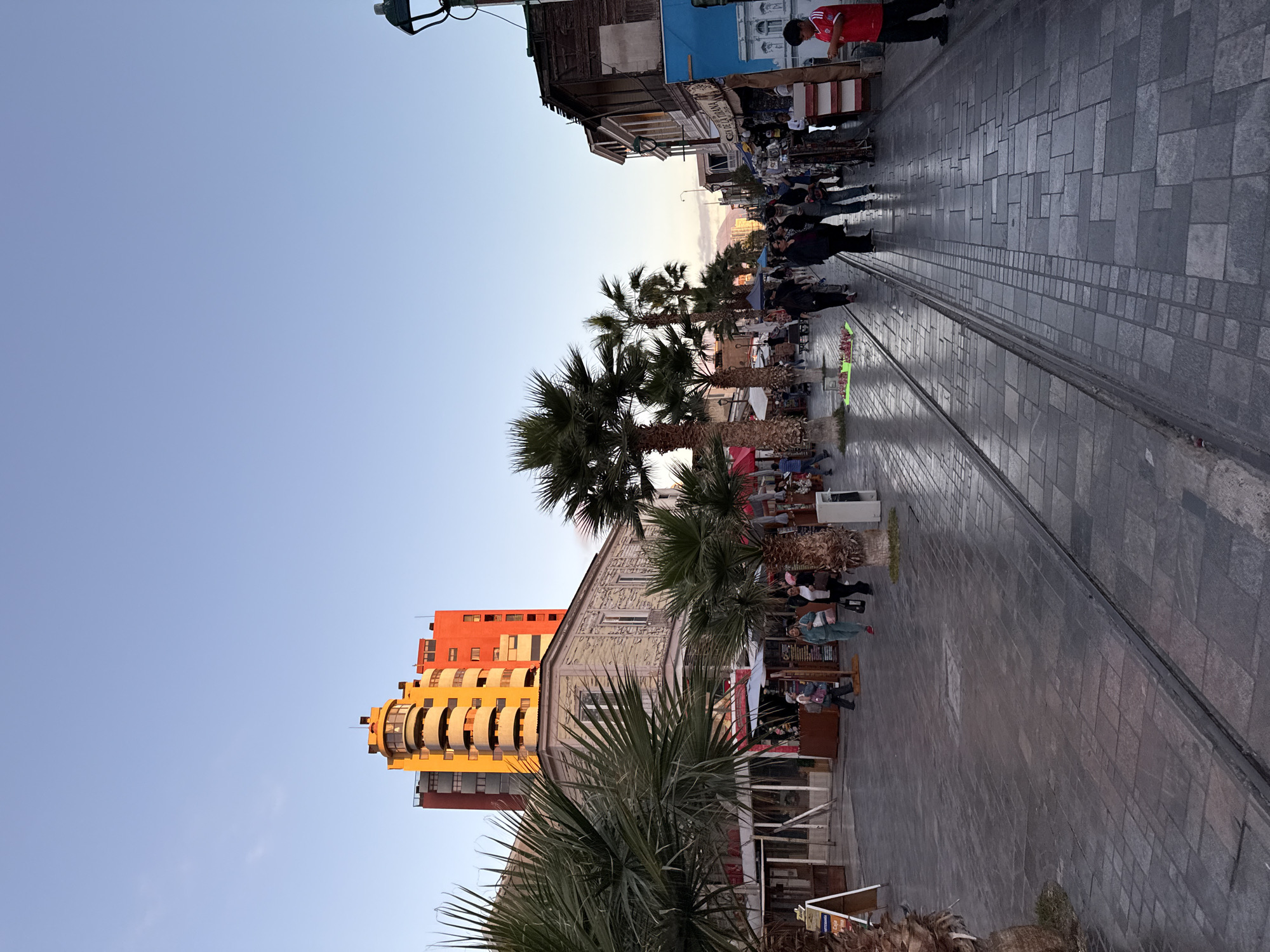 The image shows a pedestrian street in Iquique, Chile, during twilight. The street is paved with cobblestones and features tram tracks running down the center. Palm trees are planted along the sidewalks, which are lined with shops and market stalls. A colorful, multi-story building with a rounded facade stands in the background. Several people are walking along the street, browsing the shops, and interacting with each other. The sky is clear and a pale blue, indicating the end of the day. The lighting suggests a warm, pleasant evening.