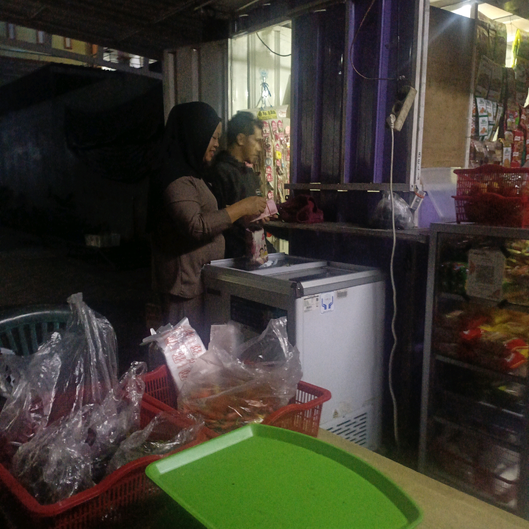 In Salatiga, Indonesia, at what appears to be a small kiosk or street-side stall, a woman wearing a dark hijab is seen interacting with a man. The woman, with her arm extended, is holding a pink object, possibly currency or a receipt, and appears to be in the process of making a transaction or receiving change. She is facing a white chest freezer, into which she is placing a small, rectangular item with images on it. To her left, a red plastic basket overflows with clear plastic bags containing various items, and another red basket is partially visible. The man, wearing a dark hoodie, stands beside her, looking towards the freezer. Behind them, shelves are stocked with packaged goods, and a power outlet is visible on a purple vertical surface. The lighting suggests it is either evening or indoors with artificial illumination. The overall scene depicts a commercial transaction occurring in a modest, possibly outdoor or semi-outdoor, retail environment.