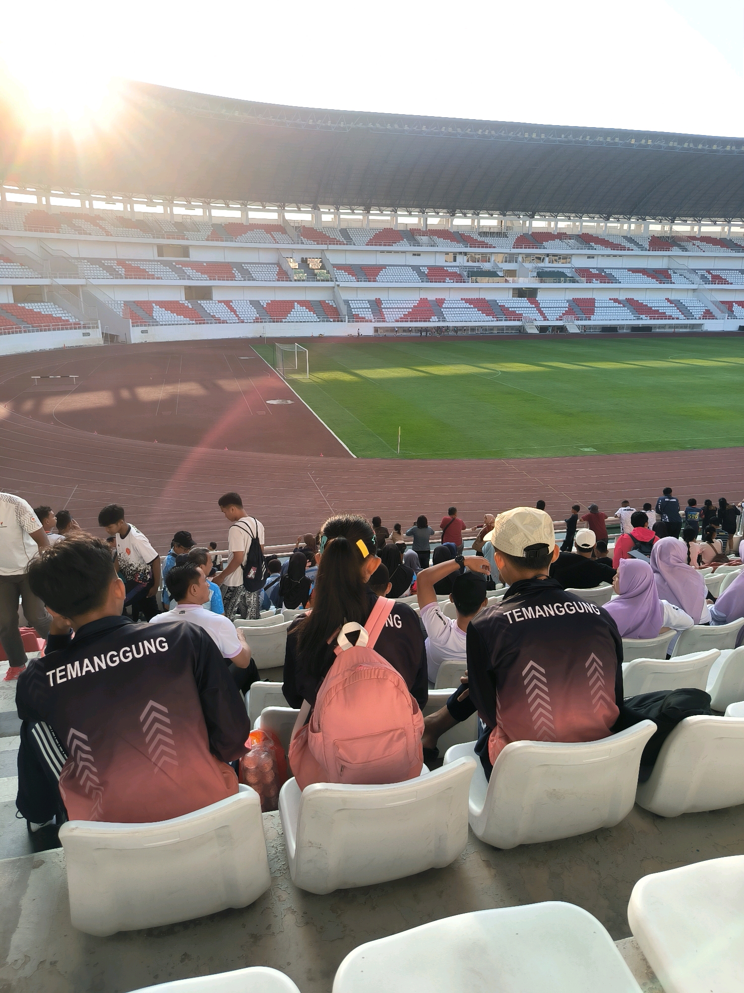 The image depicts a view from the stands of a large, open-air stadium, likely in Semarang, Indonesia, given the context. The stadium features multiple tiers of seating, with rows of white and red seats visible. The playing field, a vibrant green soccer pitch, is illuminated by the warm glow of the sun, which is low in the sky and casting long shadows. The sun's rays are also glinting off the upper stadium structure, creating a lens flare effect.
In the foreground, several individuals are seated, facing away from the camera and towards the field. They are seated on white plastic stadium chairs. Some of these individuals are wearing jackets with the word "TEMANGGUNG" printed on the back in white lettering. One person in the center foreground is wearing a pink backpack. The crowd appears to be a mix of young people and adults.
The stadium itself is a substantial structure, with a large roof covering a portion of the seating area. A running track is visible around the perimeter of the soccer field. The overall scene suggests a sports event or a gathering at a stadium, with spectators preparing to watch or observing an activity. The lighting indicates it is likely late afternoon or early evening.