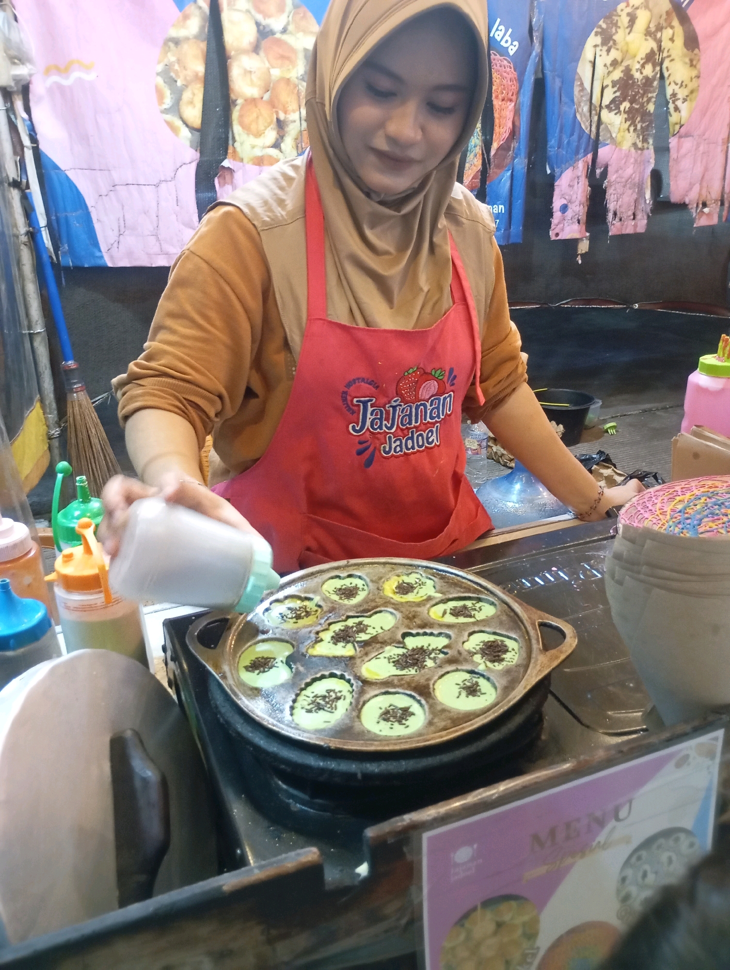 A person wearing a tan hijab and a red apron with "Jajanan Jadoel" written on it is making snacks. The person is holding a white squeeze bottle and appears to be drizzling something onto the snacks cooking in a patterned pan. The pan is on a black stove. The background shows posters with images of food and text, and there are bottles of sauces and a broom. The scene looks like a food stall in a market. The location is Babelan, Indonesia.