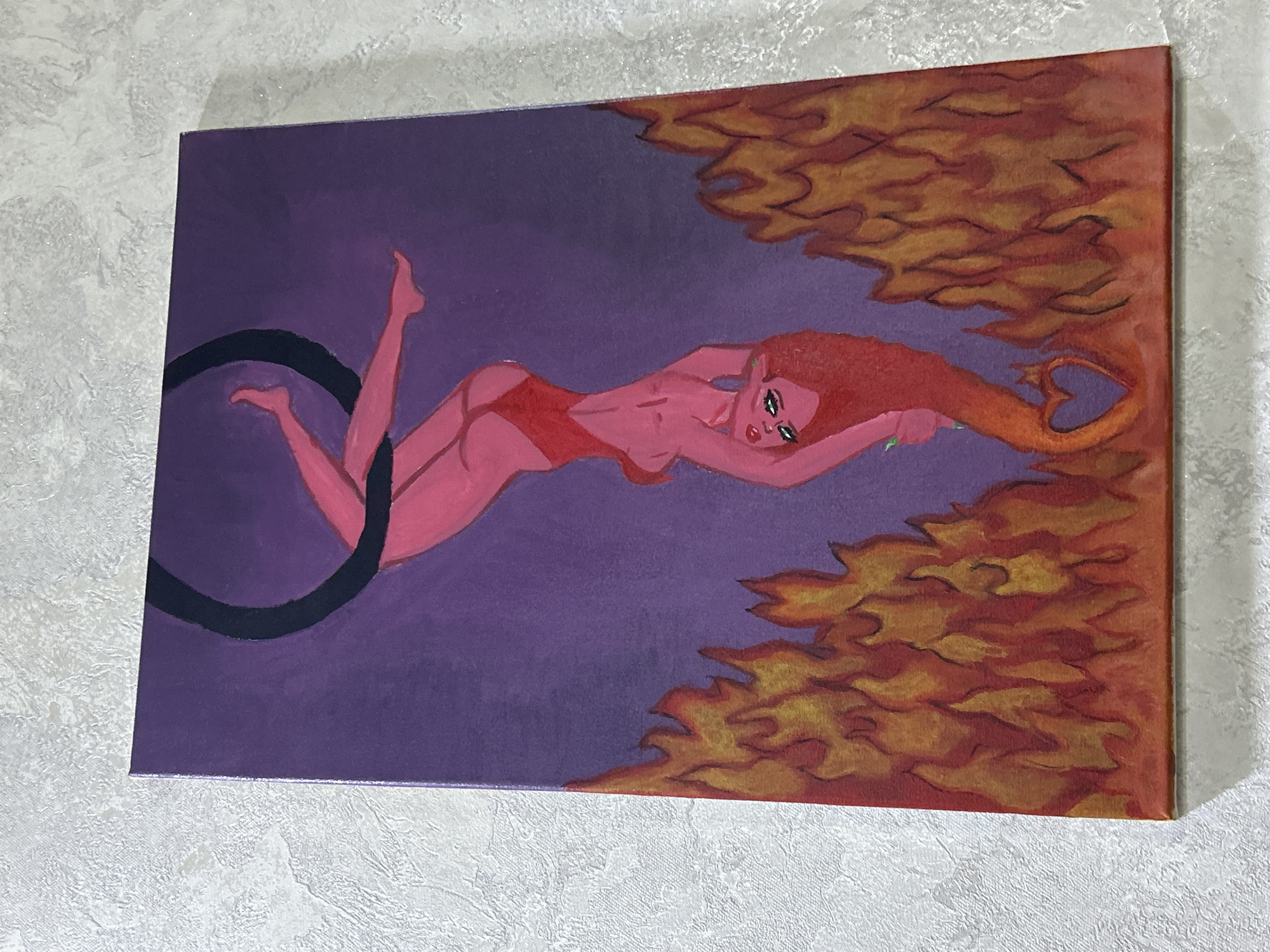 A painting on canvas depicts a stylized female figure with pink skin, red hair, and a red leotard, suspended upside down by a black hoop. The background is a gradient of purple, transitioning to orange and yellow flames at the bottom. The figure's hair flows down, forming a heart shape within the flames. The painting is hung on a textured, light-colored wall. The overall scene is artistic and symbolic, with no identifiable location in a city.