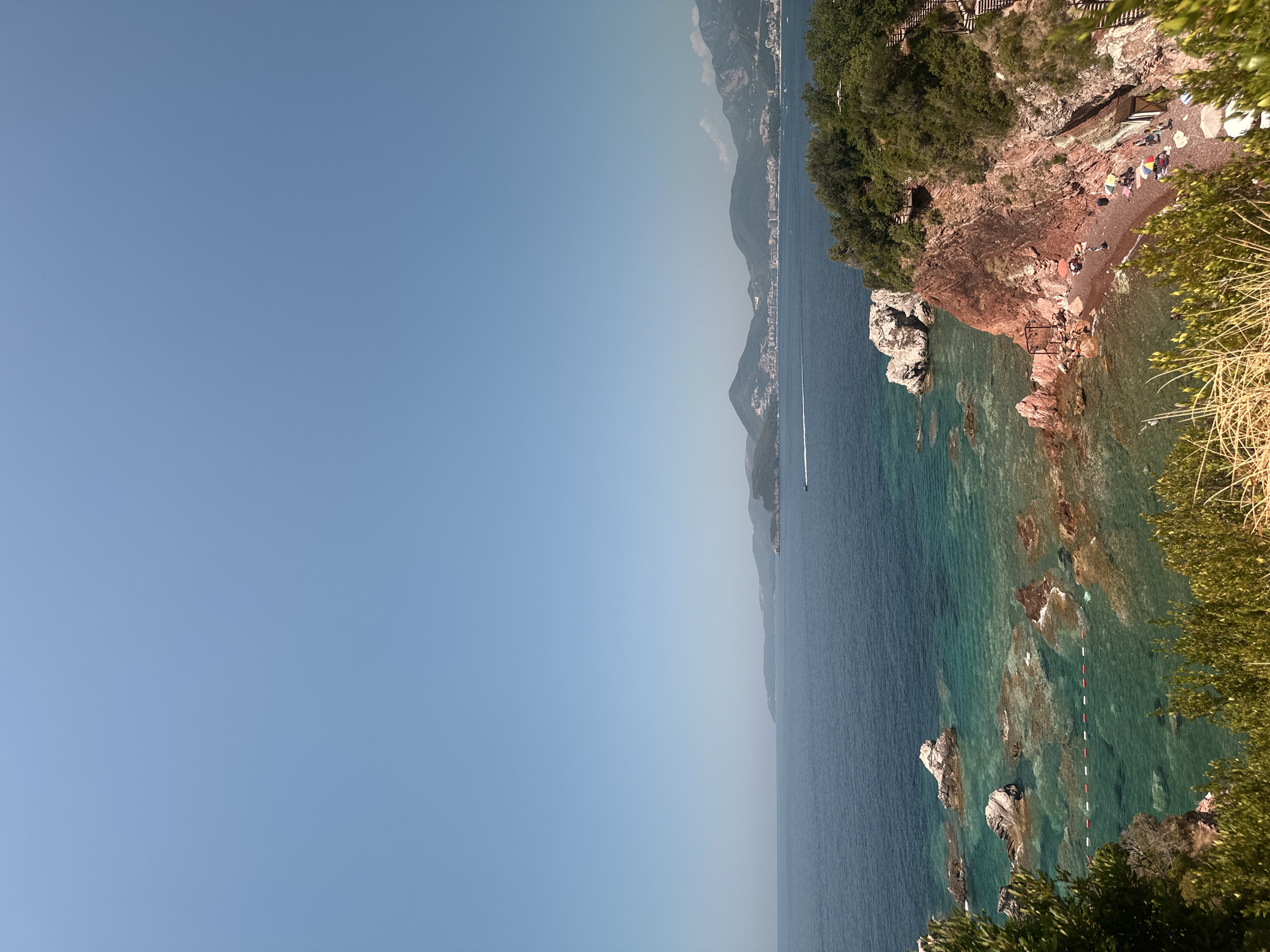 This is an aerial, high-angle, wide shot of a rocky coastline with clear, turquoise water in Sveti Stefan, Montenegro. In the foreground, there is dense green foliage. The middle ground features a rocky, red cliff face leading down to a small, secluded beach with several umbrellas and people sunbathing. The water is shallow near the shore, allowing visibility of the rocky seabed and a buouy line. Further out, the water deepens to a dark blue. In the distance, a hazy, mountainous coastline with buildings is visible under a clear, light blue sky. A small boat is also visible on the water in the mid-ground.