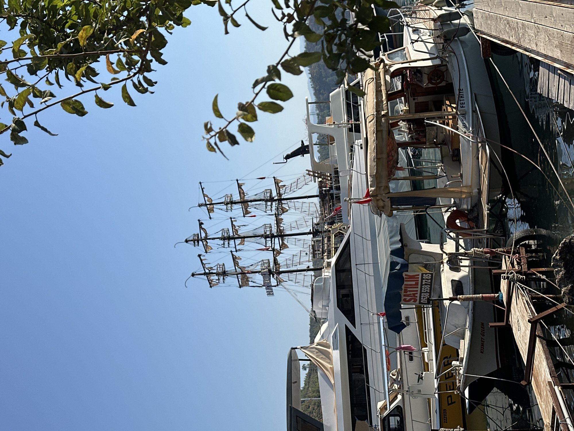 In Fethiye, Turkey, a sunny day showcases a harbor scene with several boats. A large, multi-masted schooner, reminiscent of a pirate ship, dominates the background. In the foreground, a smaller motorboat with a tan canopy is docked. To its left, another boat with a blue canopy and a "SATILIK" (For Sale) sign is visible. Further to the left, the stern of a larger white yacht is partially obscured by these boats. A wooden dock extends into the dark, reflective water. Lush green foliage from an overhanging tree frames the right side of the image, with its leaves catching the sunlight. The sky is a clear, bright blue, indicating fair weather. The overall atmosphere is serene, capturing a typical maritime setting in a coastal Turkish city.