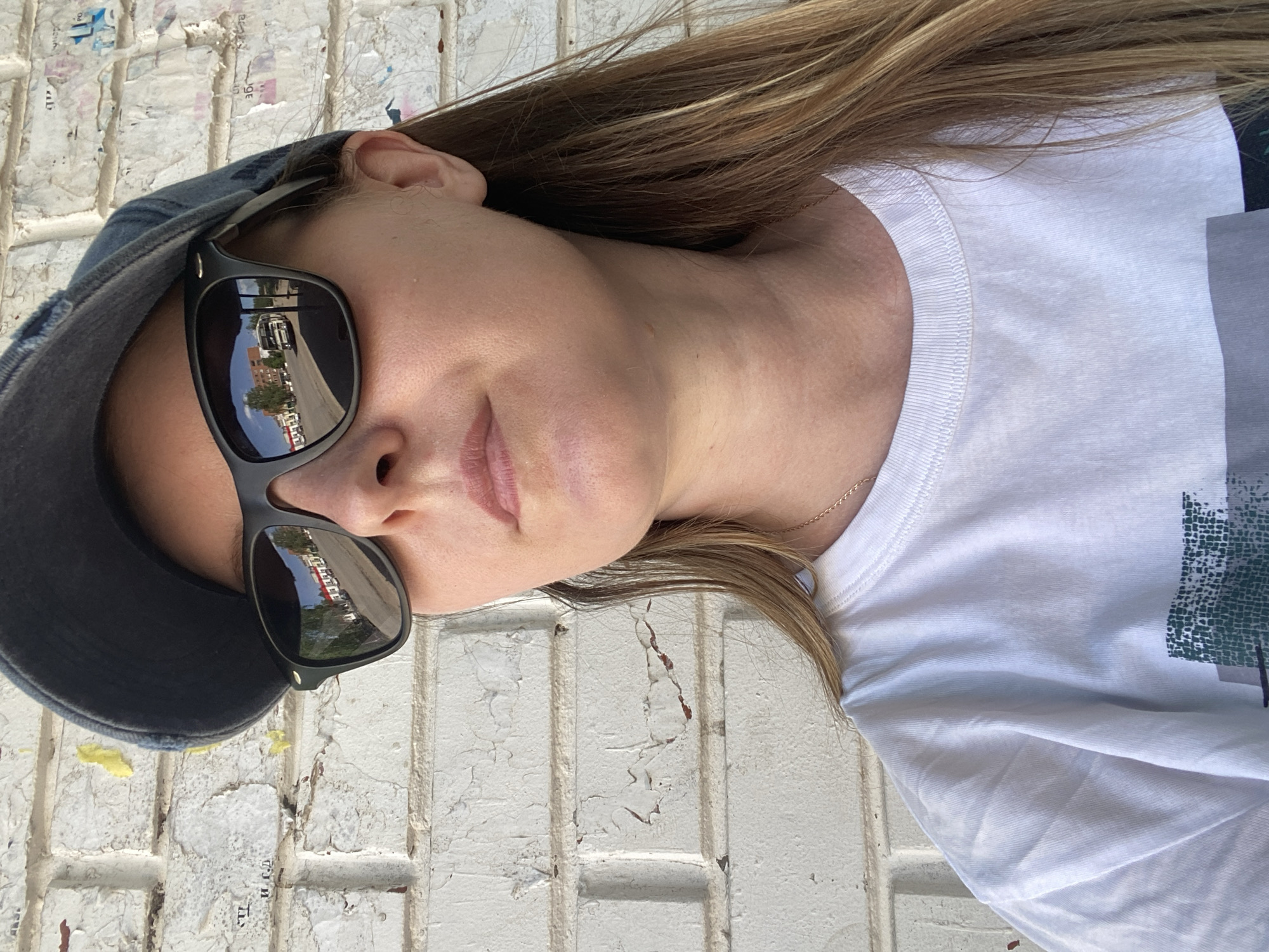 A young woman is wearing sunglasses and a baseball cap. The reflection in her sunglasses appears to depict a street in Noviy, Russia. The buildings reflected are red-brick, and there are trees visible. She is wearing a white t-shirt. The background consists of a distressed white brick wall.
